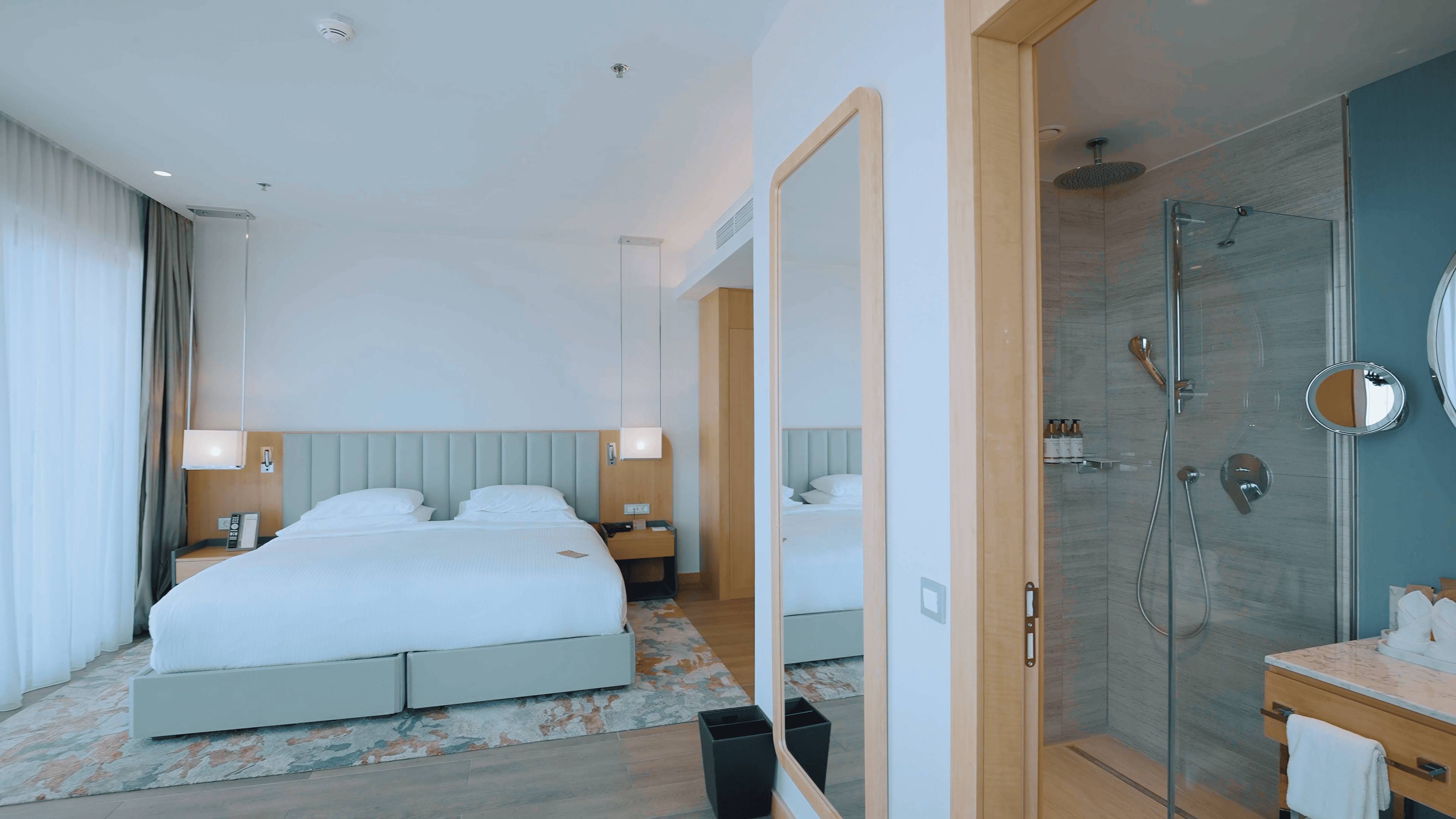 Moderna hotelska soba s bračnim krevetom i tušem otvorenog koncepta odvojenim staklenom stijenom, s ogledalom u cijeloj dužini blizu ulaza.