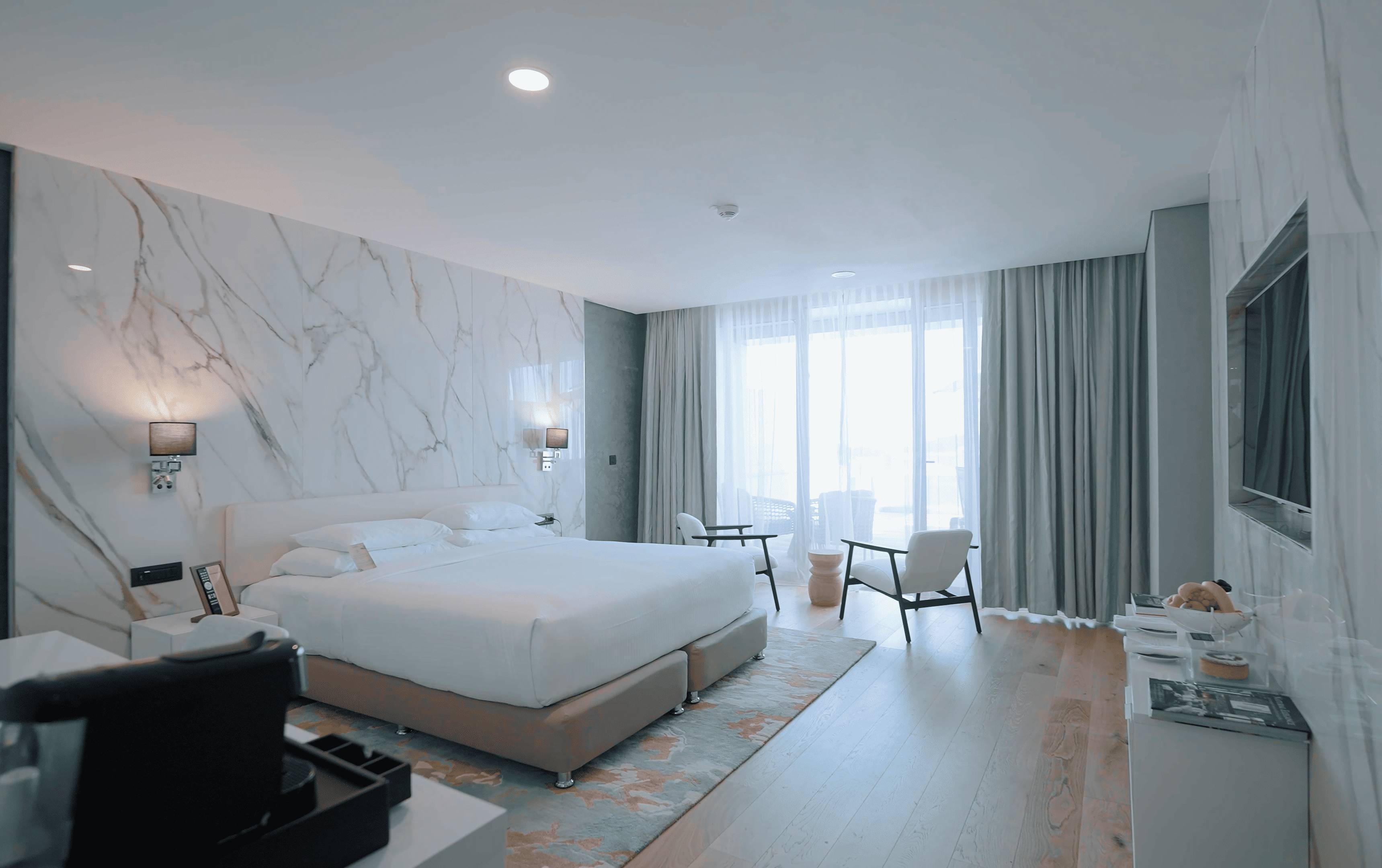 Luksuzna hotelska soba s bračnim krevetom, mramornim zidom i prostorom za sjedenje uz velike prozore s pogledom na more.