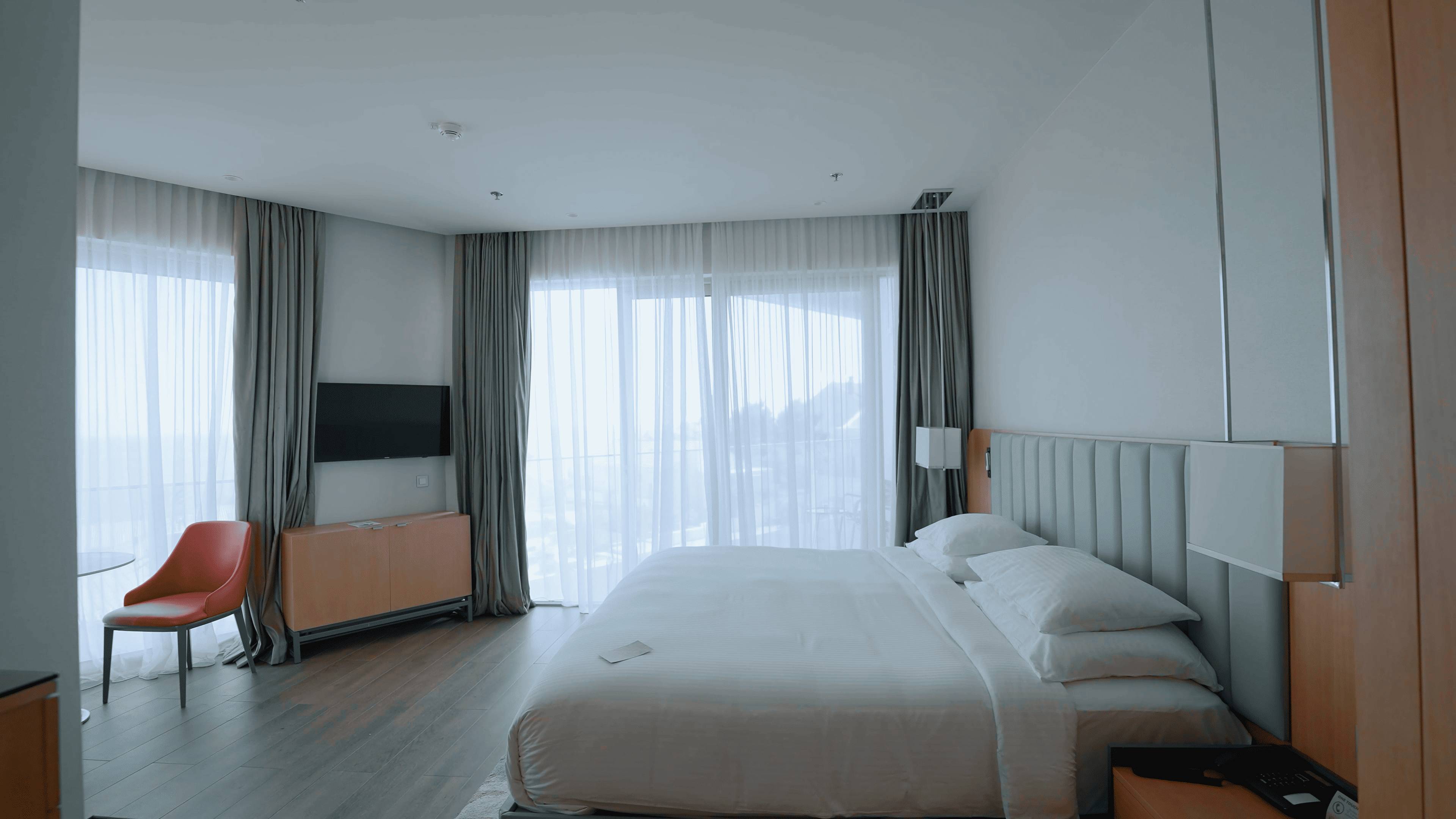 Svijetla hotelska soba s velikim bračnim krevetom, prozirnim i neprozirnim zavjesama koje prekrivaju zid od prozora, televizorom i jednom narančastom foteljom.