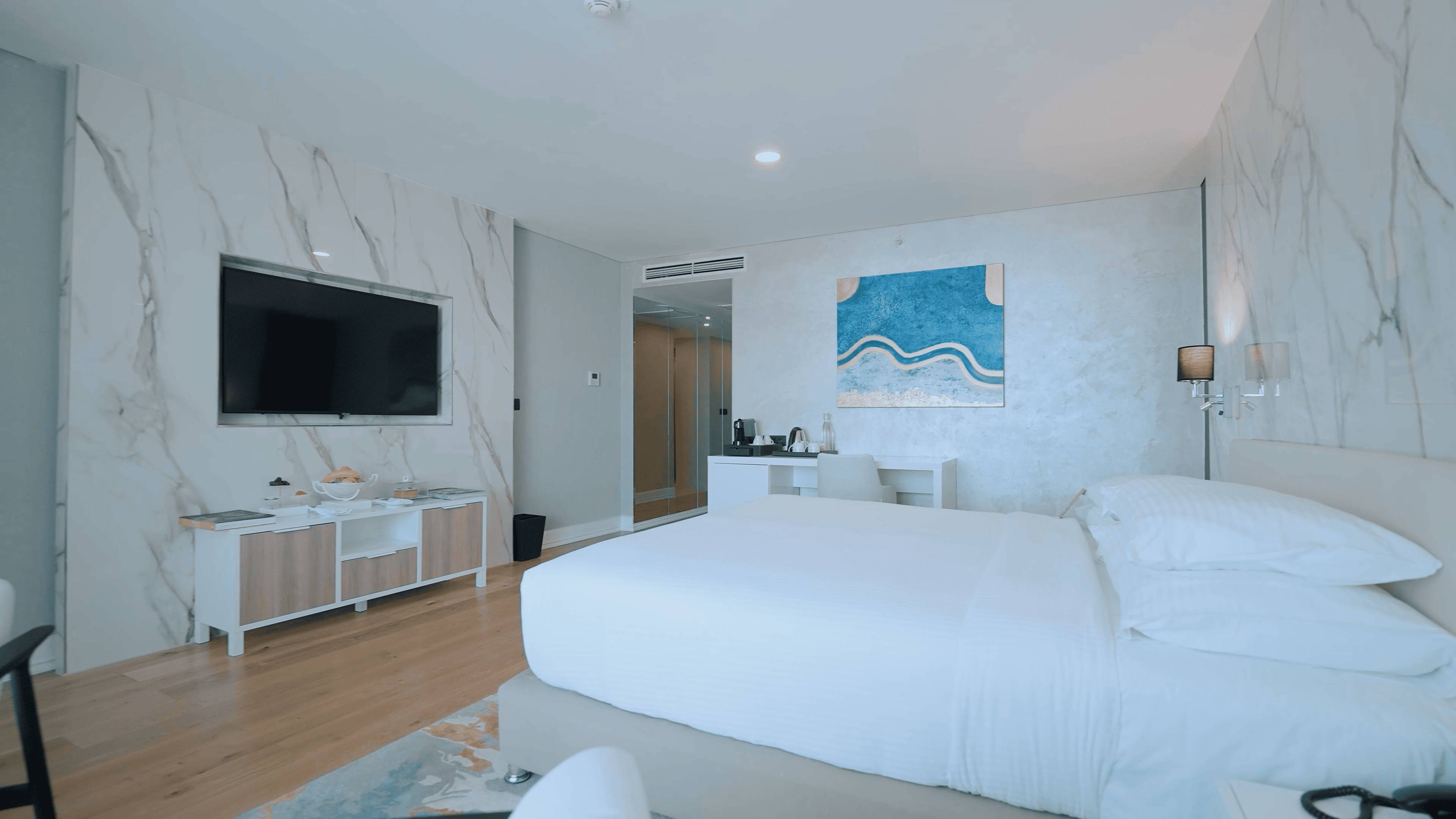 Moderna hotelska soba s bračnim krevetom, bijelim i drvenim ormarićem s TV-om i plavom apstraktnom slikom iznad radnog stola