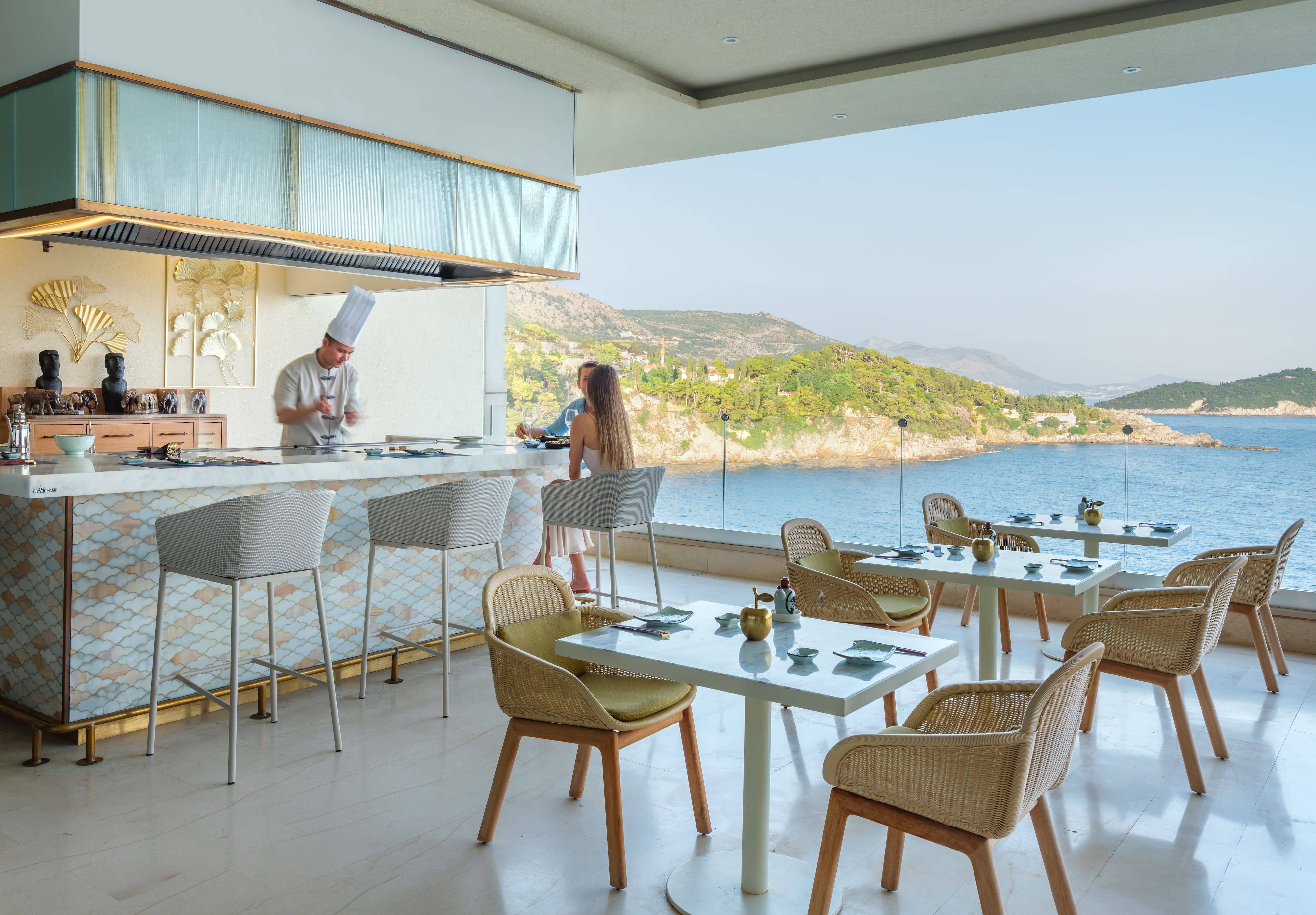 An image of the Umi Teppanyaki restaurant at the Rixos Premium Dubrovnik. The open-air restaurant features a Teppanyaki bar on the left, where a chef in a white toque is preparing food, and a woman is seated at the bar. Several dining tables with light-colored wicker chairs are set on the right. The restaurant has a floor-to-ceiling opening with a glass railing, providing a breathtaking view of the blue Adriatic Sea and the lush green coastline of Dubrovnik.