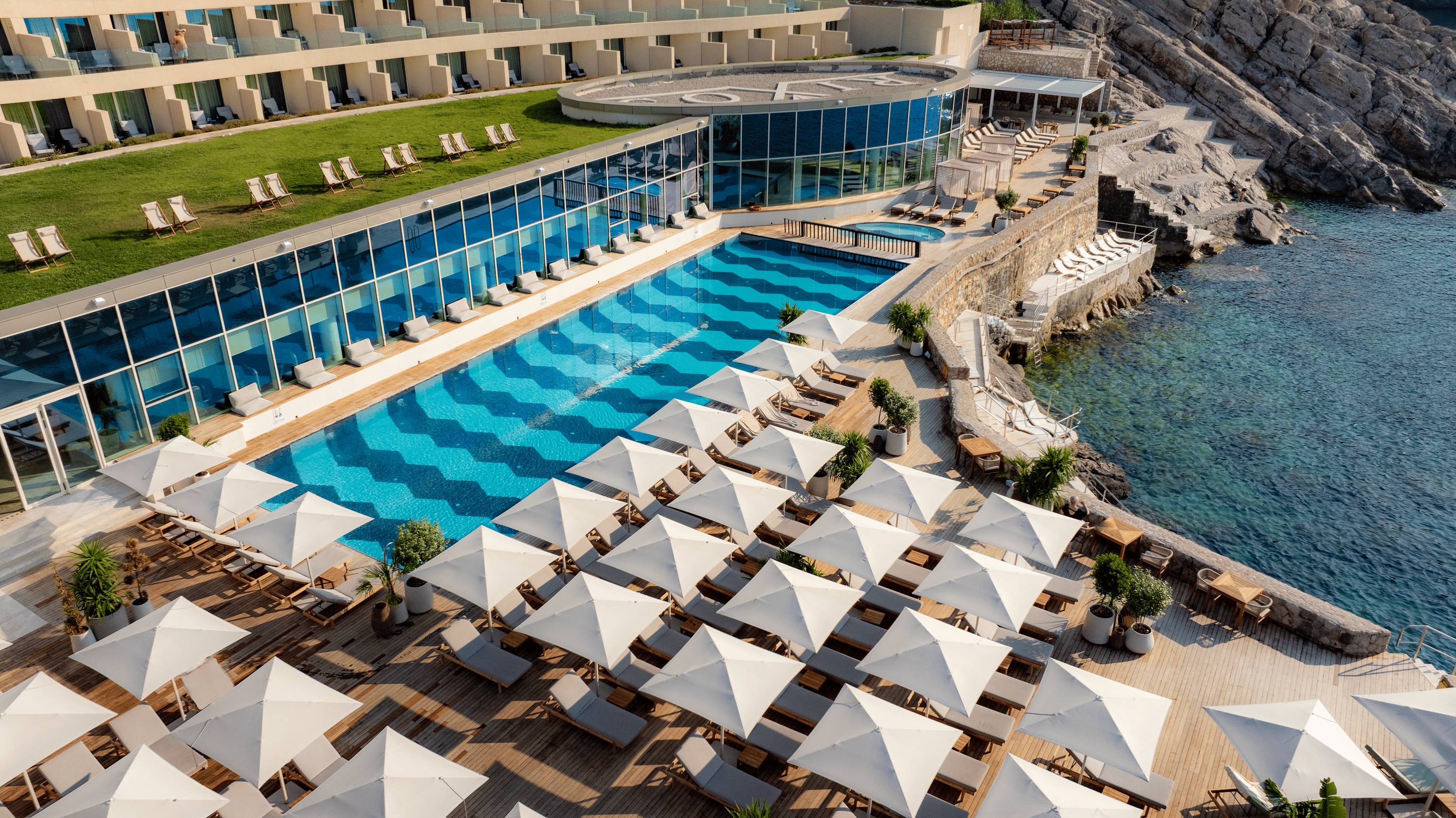 A high-angle view of the Mykorini Beach area at the Rixos Premium Dubrovnik. The area features a large, rectangular swimming pool with a distinctive blue and white wave pattern on the bottom. The pool deck is covered with numerous white square parasols over sun loungers. The modern hotel building is visible on the left, and the deck extends down to the rocky coastline and the sea on the right.