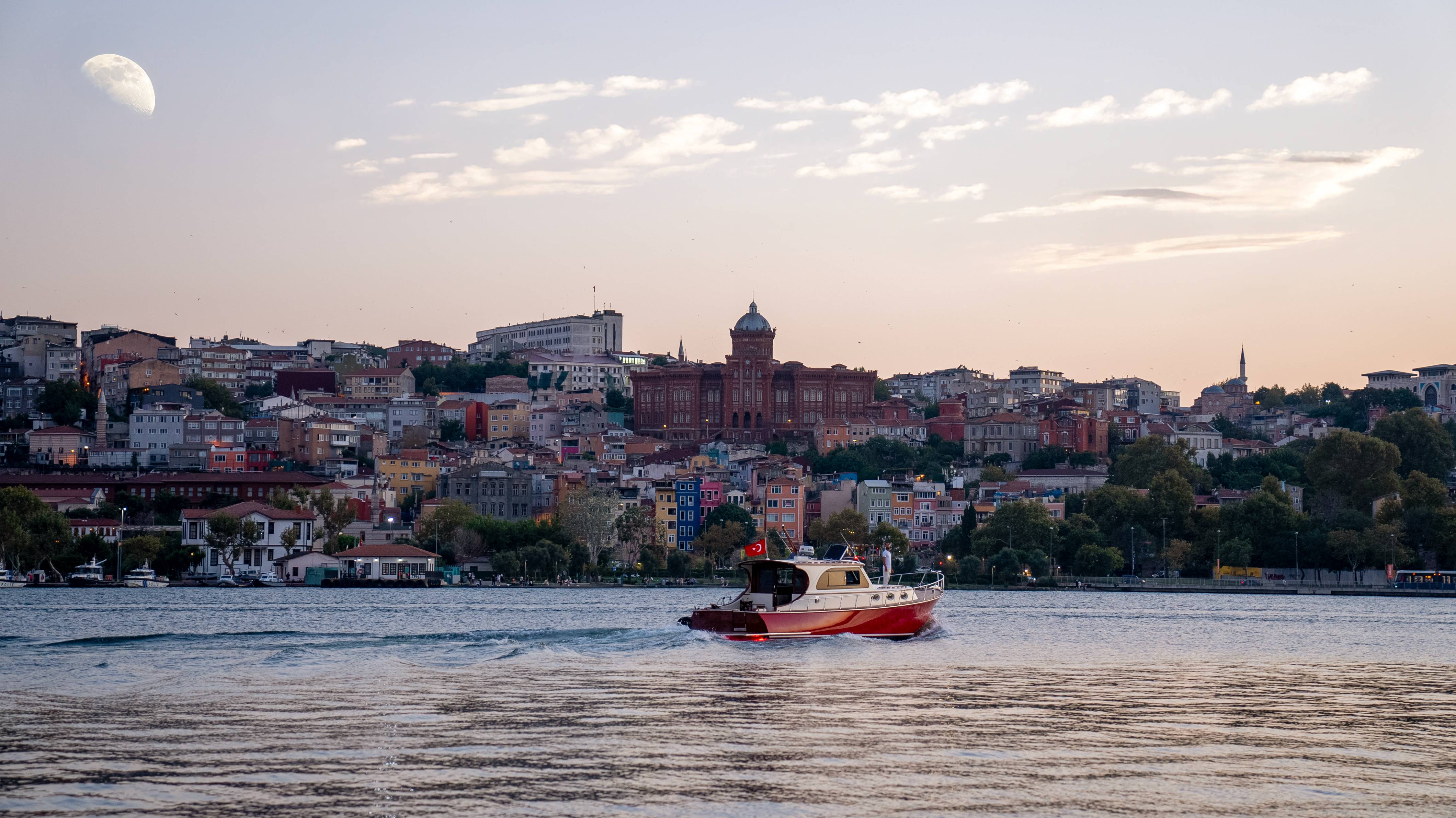 Malowniczy widok na łódź płynącą wzdłuż Złotego Rogu podczas wycieczki z przewodnikiem Rixos Tersane Istanbul Fener-Balat, z kolorowymi domami na wzgórzach i zabytkową architekturą w tle pod wschodzącym księżycem.