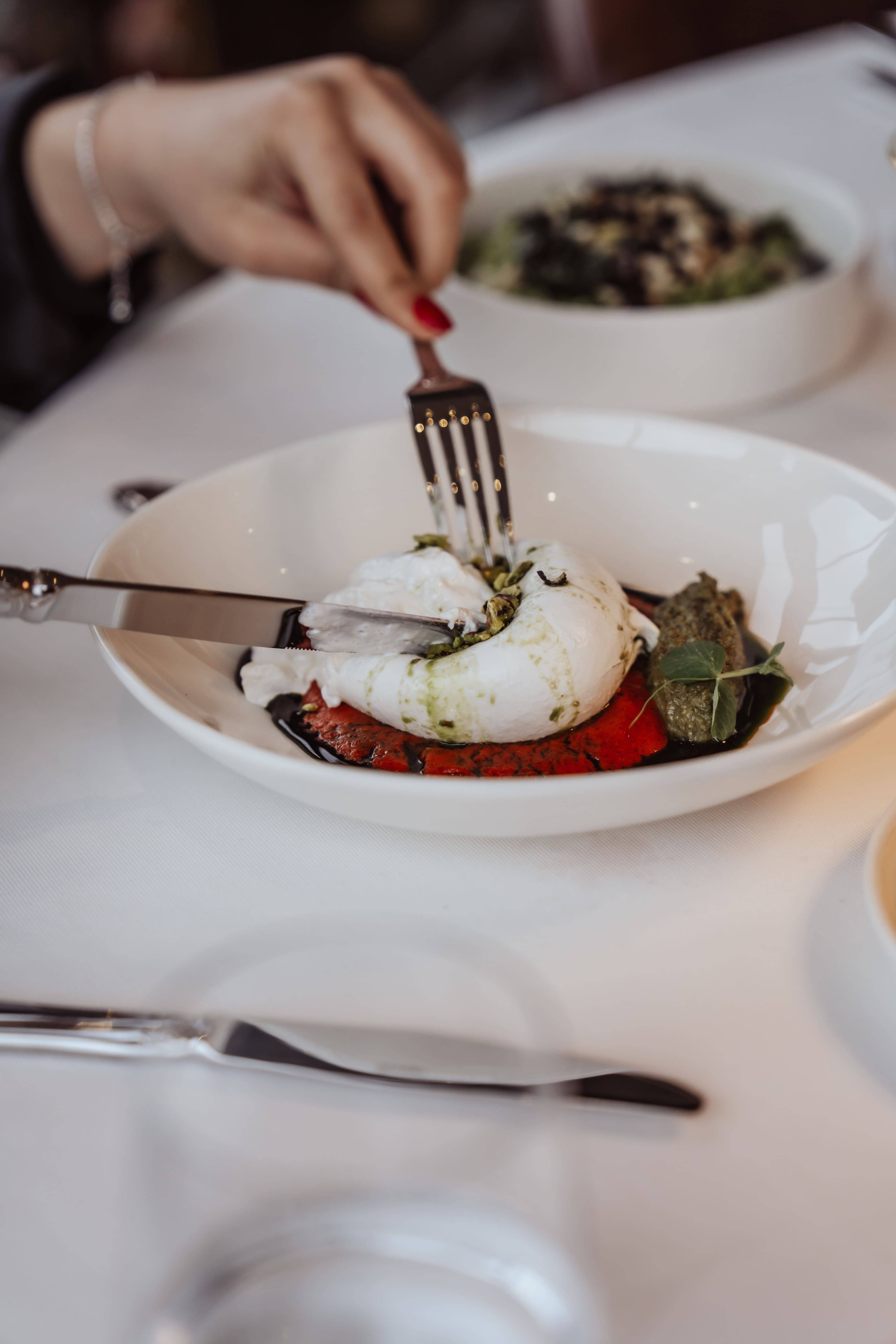 Krupni plan ruke s crvenim lakom za nokte kako reže kuglicu sira Burrata posluženu s balzamičnom glazurom i pestom na crvenoj podlozi u restoranu Vetiver, Rixos Pera Istanbul.