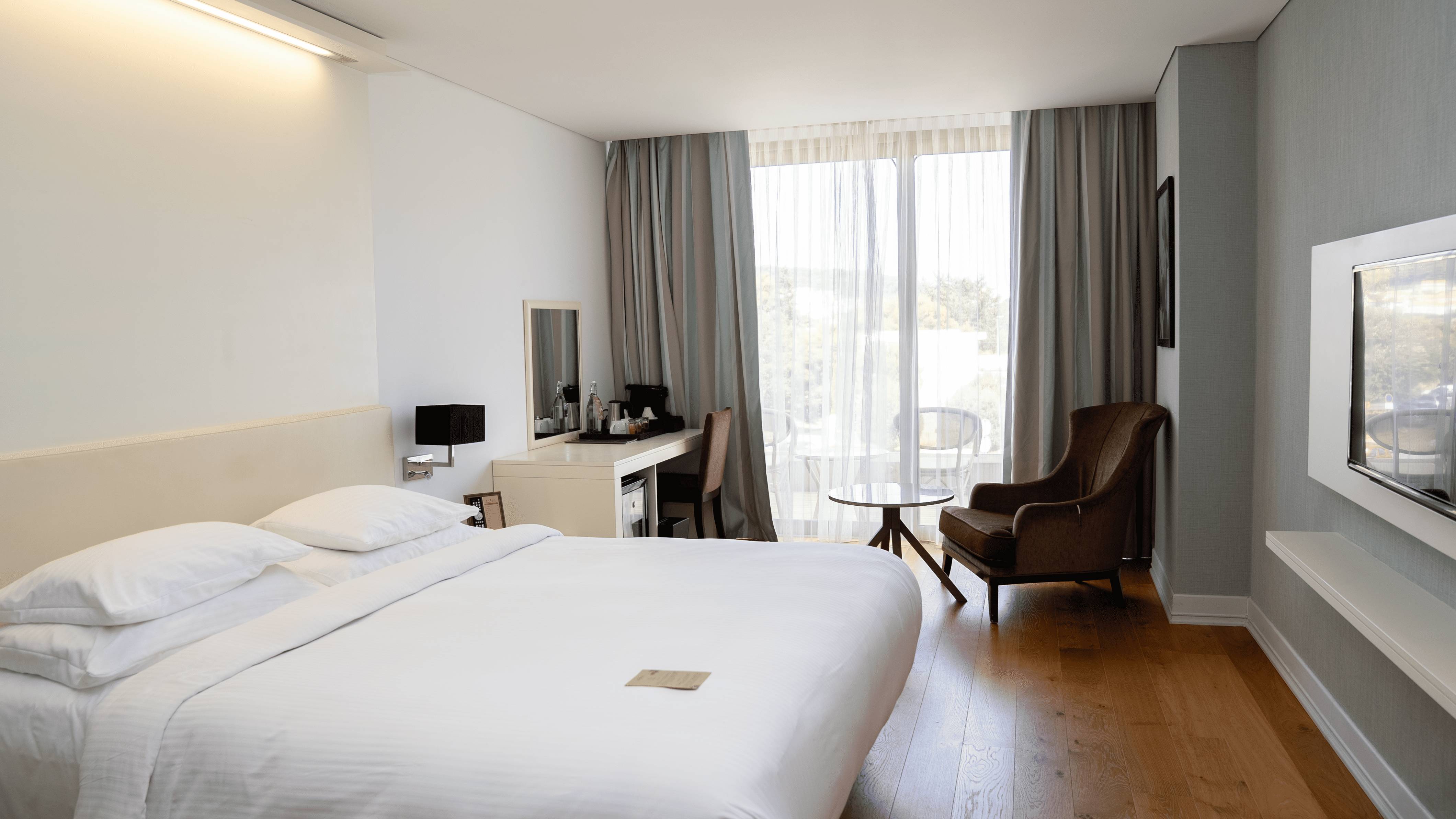 Hotelska spavaća soba s bračnim krevetom, drvenim podom, prozirnim zavjesama, pisaćim stolom i tapeciranom naslonjačem blizu balkona.