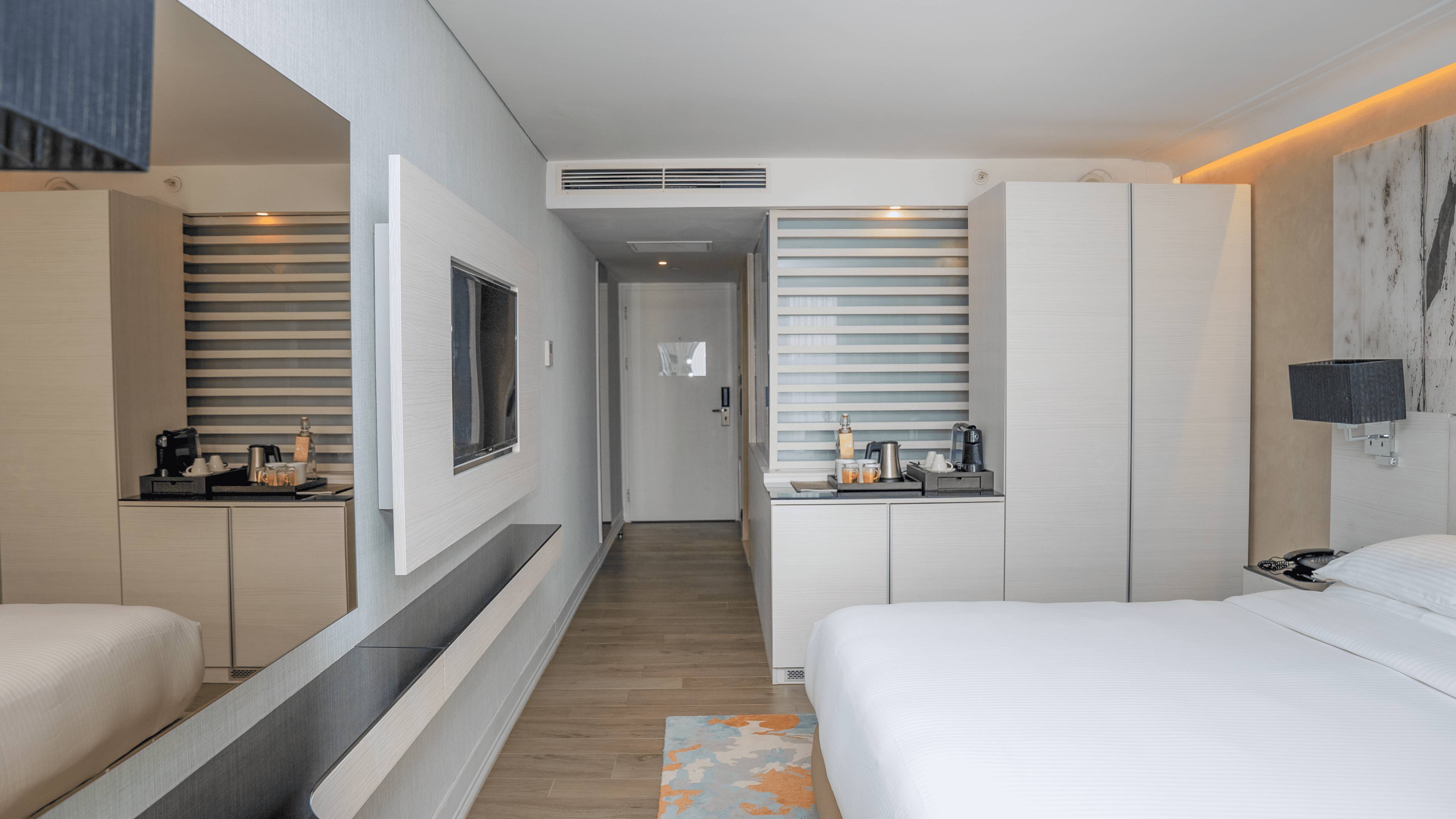 Hotelska soba s dva odvojena kreveta, velikim zidnim televizorom i kliznim staklenim vratima koja vode na balkon s čistim pogledom na more.