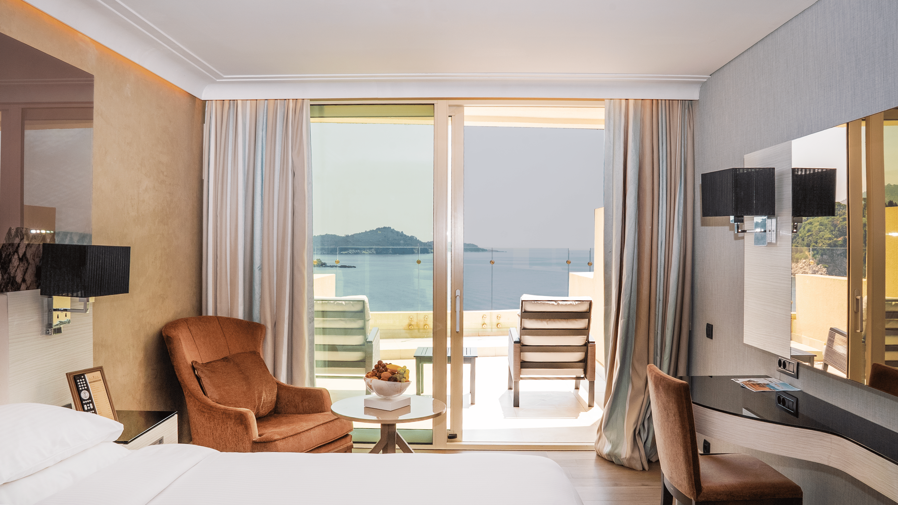 Hotelska soba s bračnim krevetom, crvenom radnom stolicom, dvije fotelje i kliznim staklenim vratima koja vode na travnatu terasu s čistim pogledom na more.