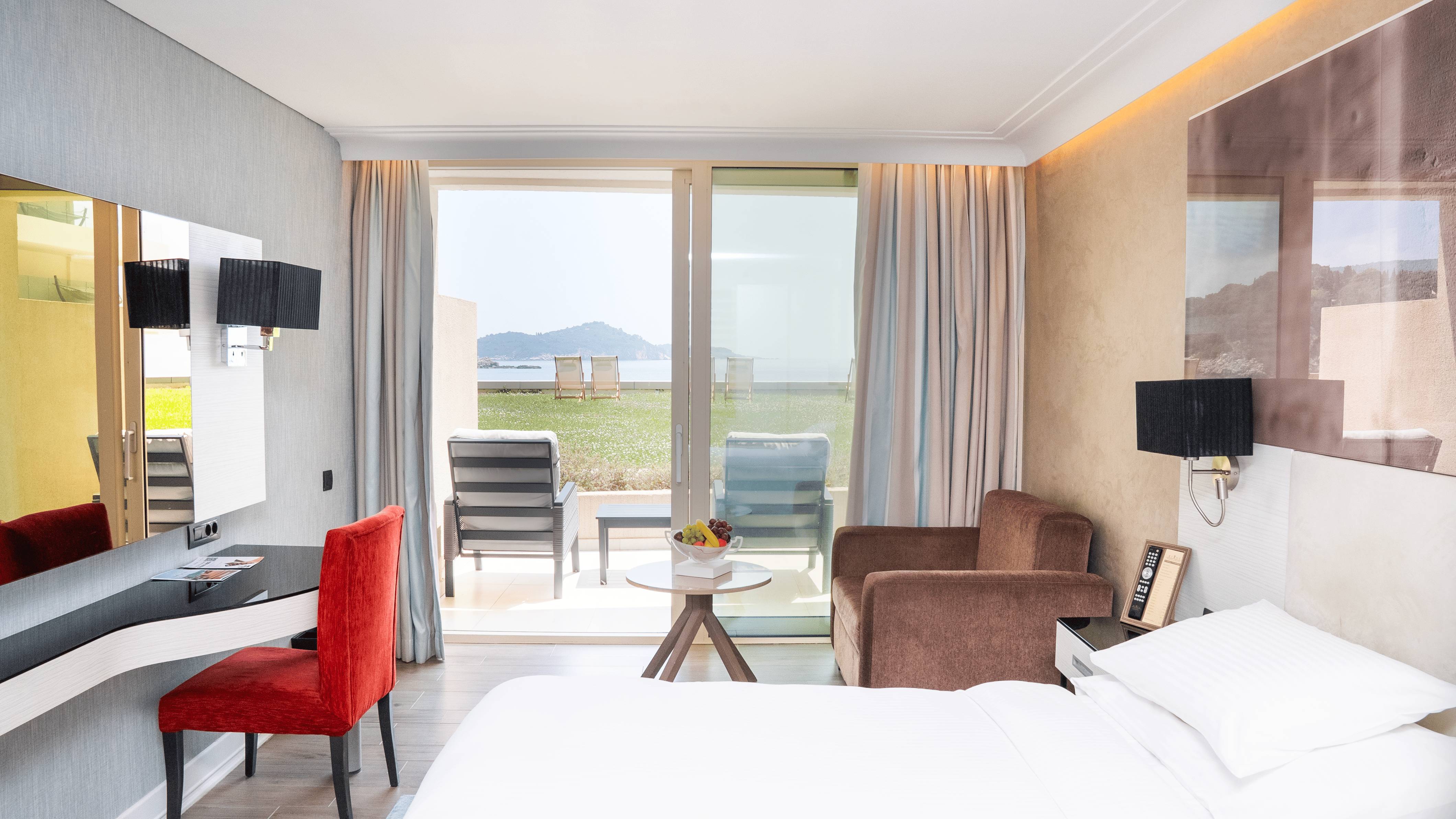 Hotelska soba s bračnim krevetom, crvenom radnom stolicom, dvije fotelje i kliznim staklenim vratima koja vode na travnatu terasu s pogledom na more
