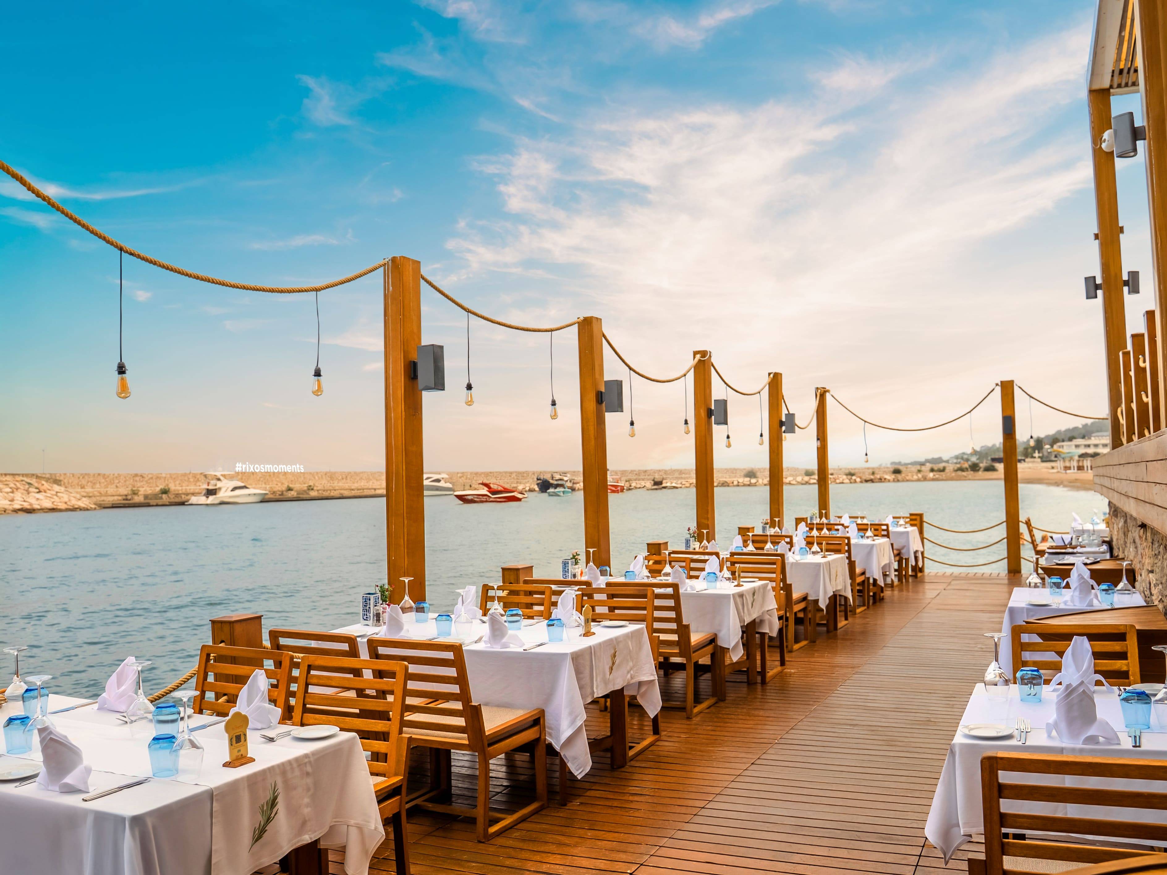 Elegant seaside dining setup at La Rosetta À La Carte Restaurant, Rixos Sungate, featuring wooden tables with white linens overlooking the marina and yachts under a bright blue sky.