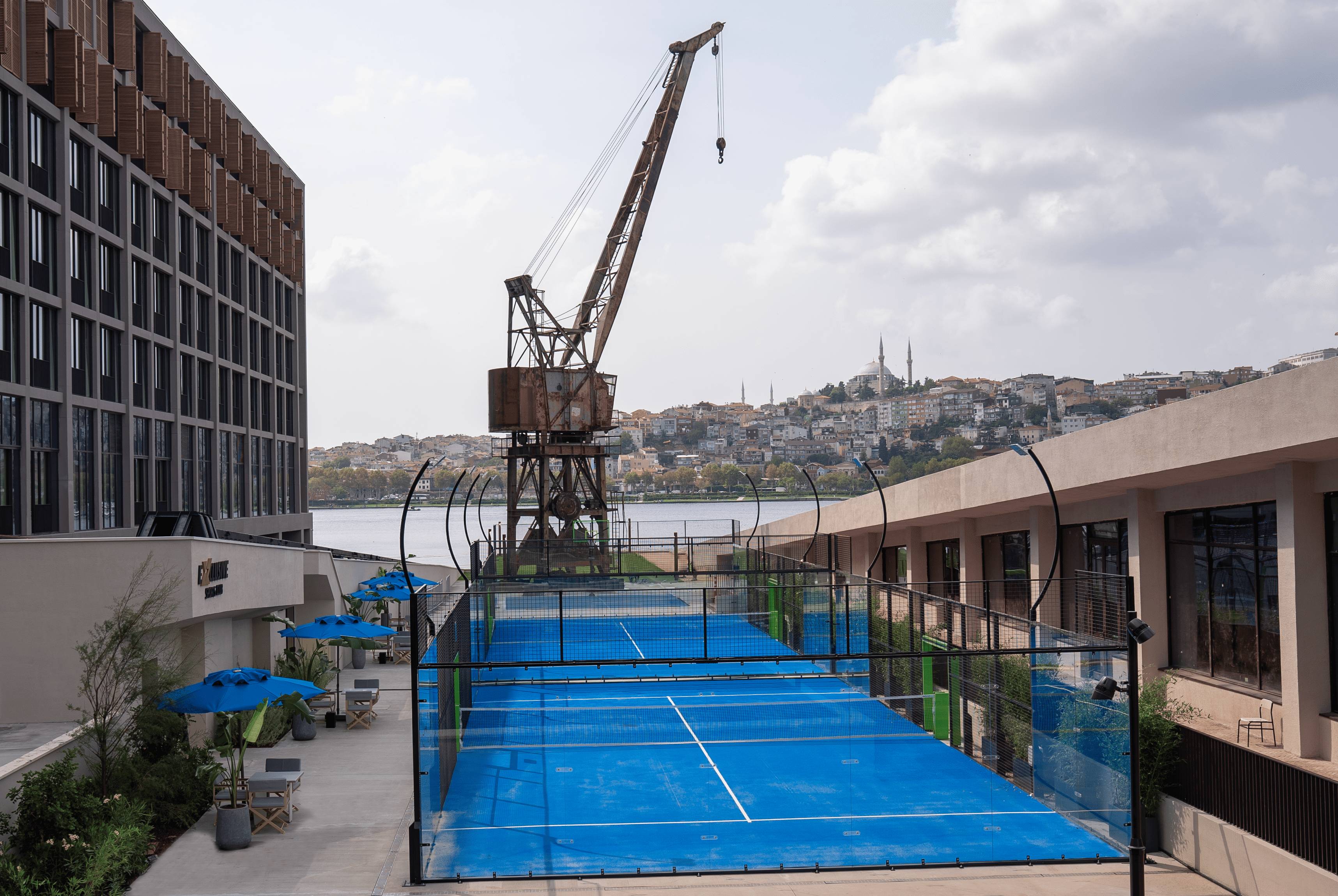Korty tenisowe Blue Padel w hotelu Rixos Tersane Istanbul z widokiem na Złoty Róg, historyczną konstrukcję dźwigu i panoramę miasta w tle.
