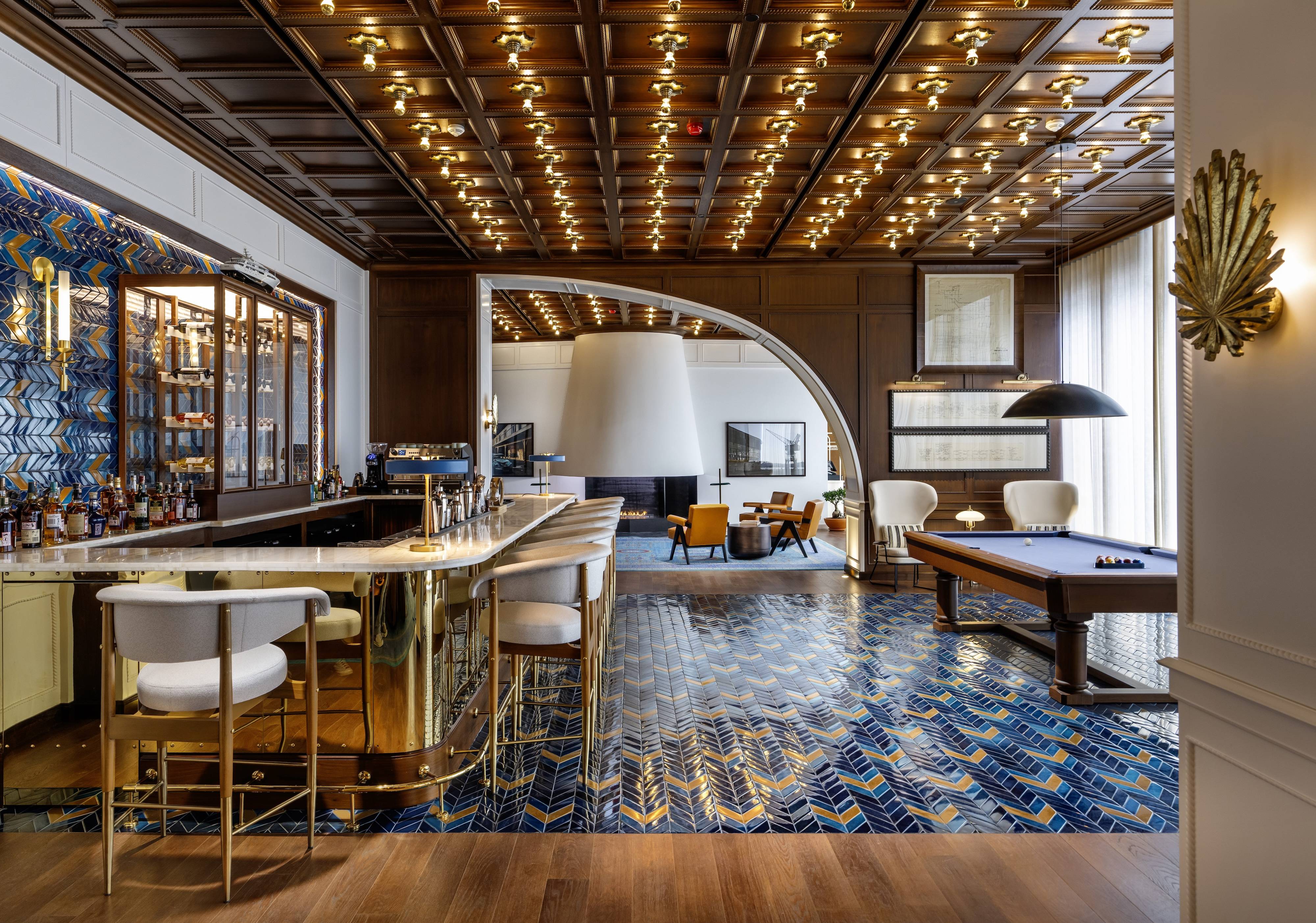 Luksusowy salon w hotelu Rixos Tersane Istanbul wyposażony jest w mosiężny bar z białymi stołkami, podłogę wyłożoną niebieskimi wzorzystymi kafelkami, sufit wyłożony drewnianymi panelami z wieloma lampami, stół bilardowy i nowoczesny kominek z miejscami do siedzenia w klubie Gowden.