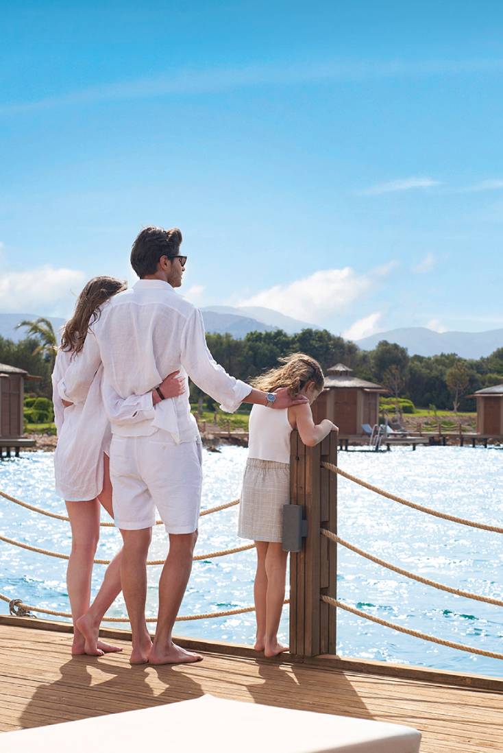 A family of three, dressed in light summer clothes, stands on a wooden deck at Rixos Park Belek, overlooking a body of water, with small wooden cabins and trees visible in the background under a bright blue sky.