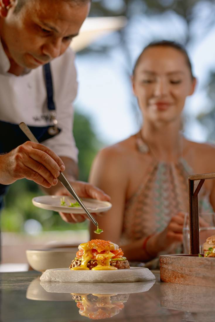 A chef at Rixos Park Belek garnishes a dish with green herbs using tweezers, while a woman in a halter dress watches and smiles in the bright outdoor setting—an inviting moment with Infinia Vacation Club.