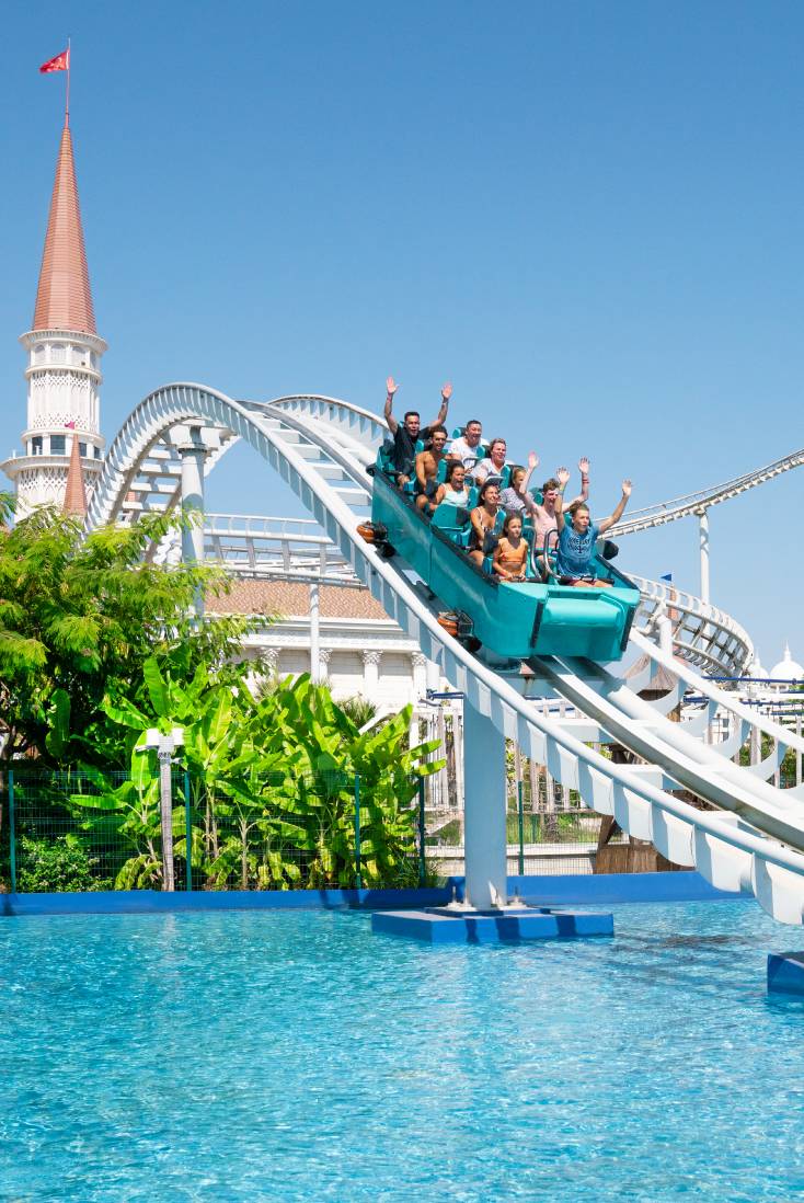 A group of people ride a roller coaster at The Land of Legends Theme Park over a pool of blue water, raising their hands in excitement, with greenery, a white building, and a pointed tower in the background under a clear blue sky.