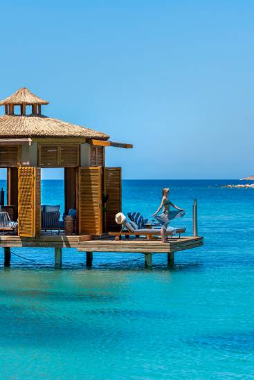A wooden cabana stands over clear turquoise water under a blue sky. At Rixos Park Belek, a woman in a sunhat and dress balances on one leg by a lounge chair, enjoying the ocean view—an unforgettable Infinia Vacation Club escape