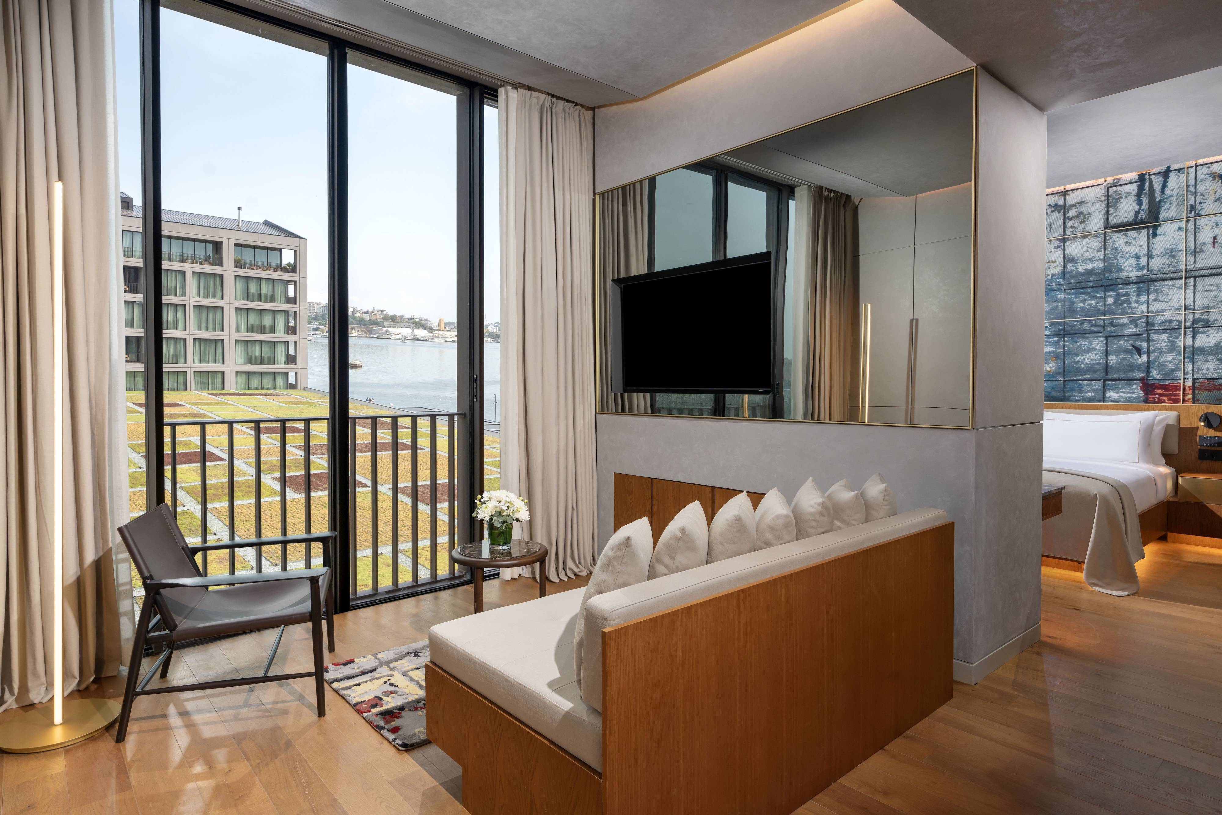 Rixos Tersane Istanbul Junior Suite Partial Sea View to nowoczesny pokój hotelowy z dużymi oknami z widokiem na wodę i pobliskie budynki, wyposażony w drewnianą sofę, telewizor na ścianie, świeże kwiaty i schludnie pościelone łóżko.