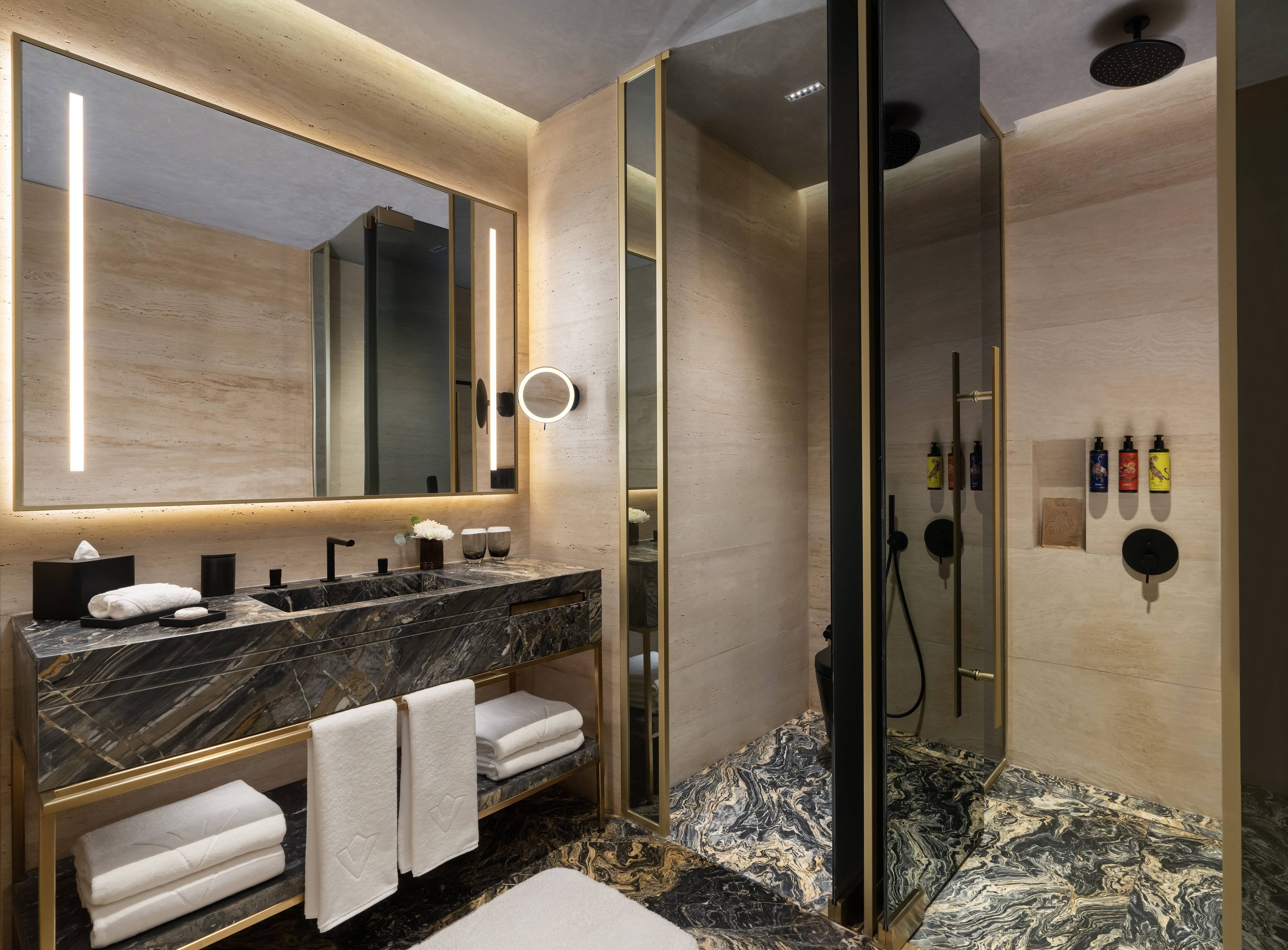 A modern bathroom in the Junior Suite Partial Sea View at Rixos Tersane Istanbul features a large illuminated mirror, double sink with black marble countertop, gold accents, white towels, a glass-enclosed shower, and beige stone walls and floors.