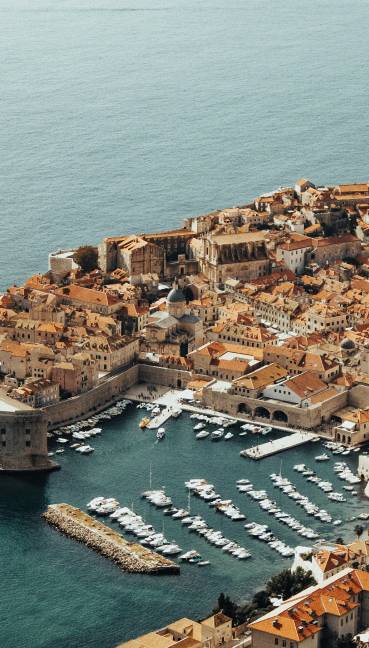 Aerial view of a historic coastal city with orange-roofed buildings, stone walls, and a bustling harbor near the blue sea, with Rixos Premium Dubrovnik adding modern luxury to the timeless landscape.