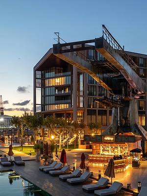 Nowoczesny hotel Rixos Tersane Istanbul z dużymi szklanymi oknami i unikalną architekturą industrialną wychodzi na basen otoczony leżakami i parasolami o zachodzie słońca. Tekst brzmi: Last Minute – Skorzystaj teraz z okazji, aby uciec do Stambułu.