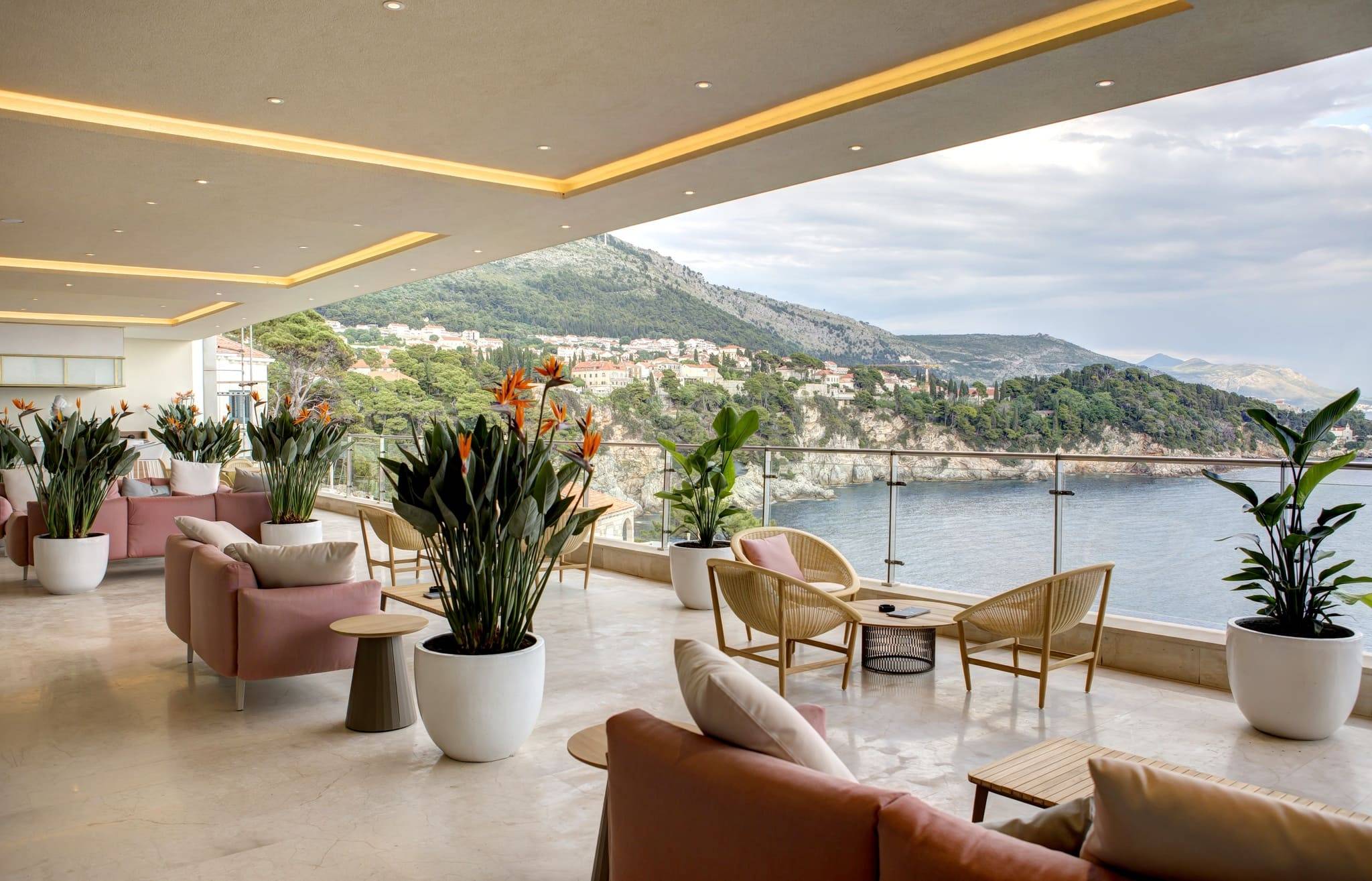 A view of the outdoor seating area at the Libertas Terrace & Lobby Bar at Rixos Premium Dubrovnik. The modern, covered terrace features light pink sofas and natural wicker chairs and tables, interspersed with tall planters holding Bird of Paradise and other lush plants. The space overlooks the Adriatic Sea and the rugged coastline with houses, visible through a clear glass railing.