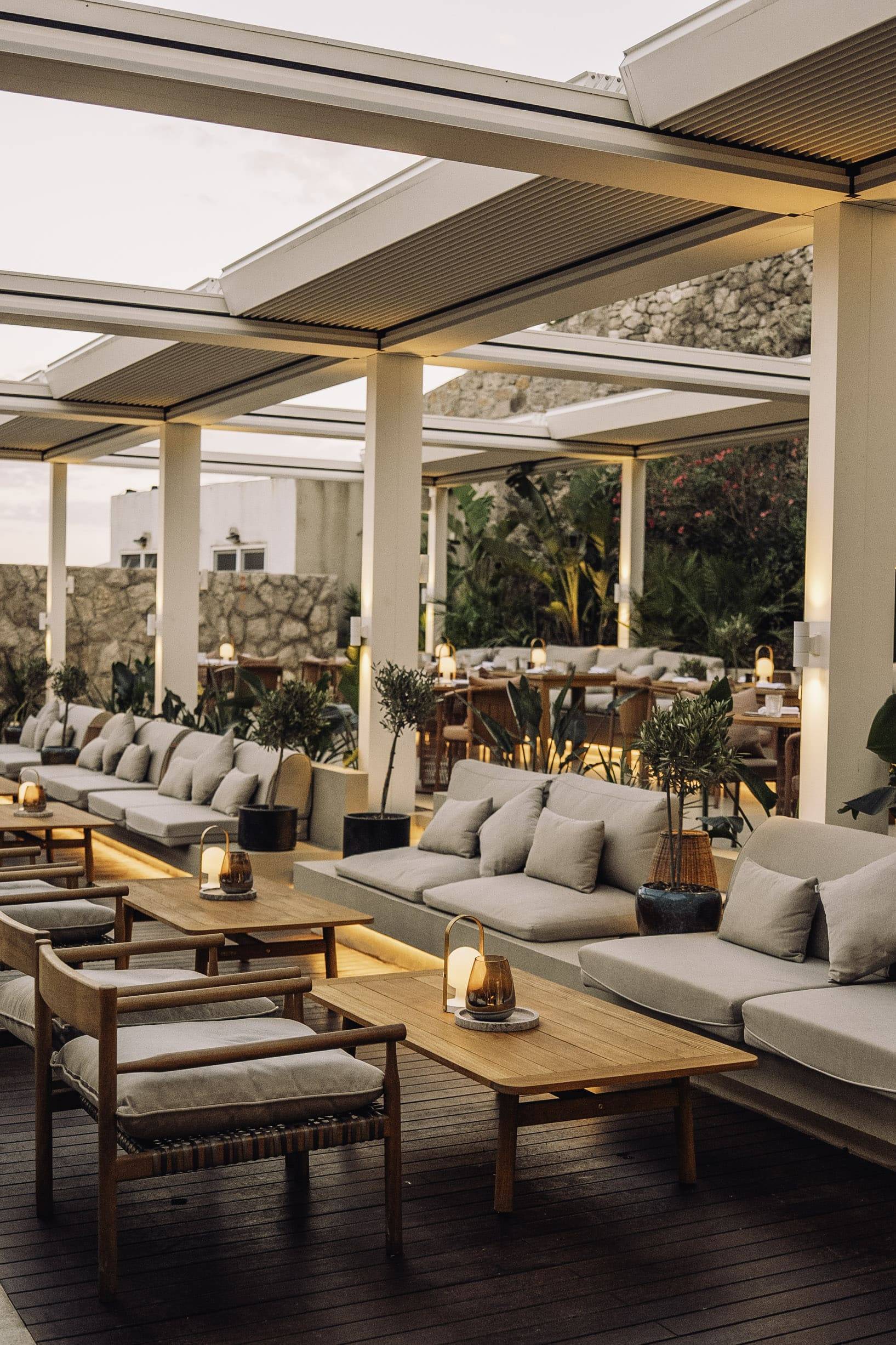 A stylish outdoor lounge area at the Mykorini Restaurant at Rixos Premium Dubrovnik. The space features long, low modular sofas and wooden armchairs with light gray cushions, arranged around low wooden tables. Small, warm lantern lights sit on the tables, creating a cozy evening atmosphere under a modern, slatted pergola roof. Lush greenery and stonework are visible in the background.