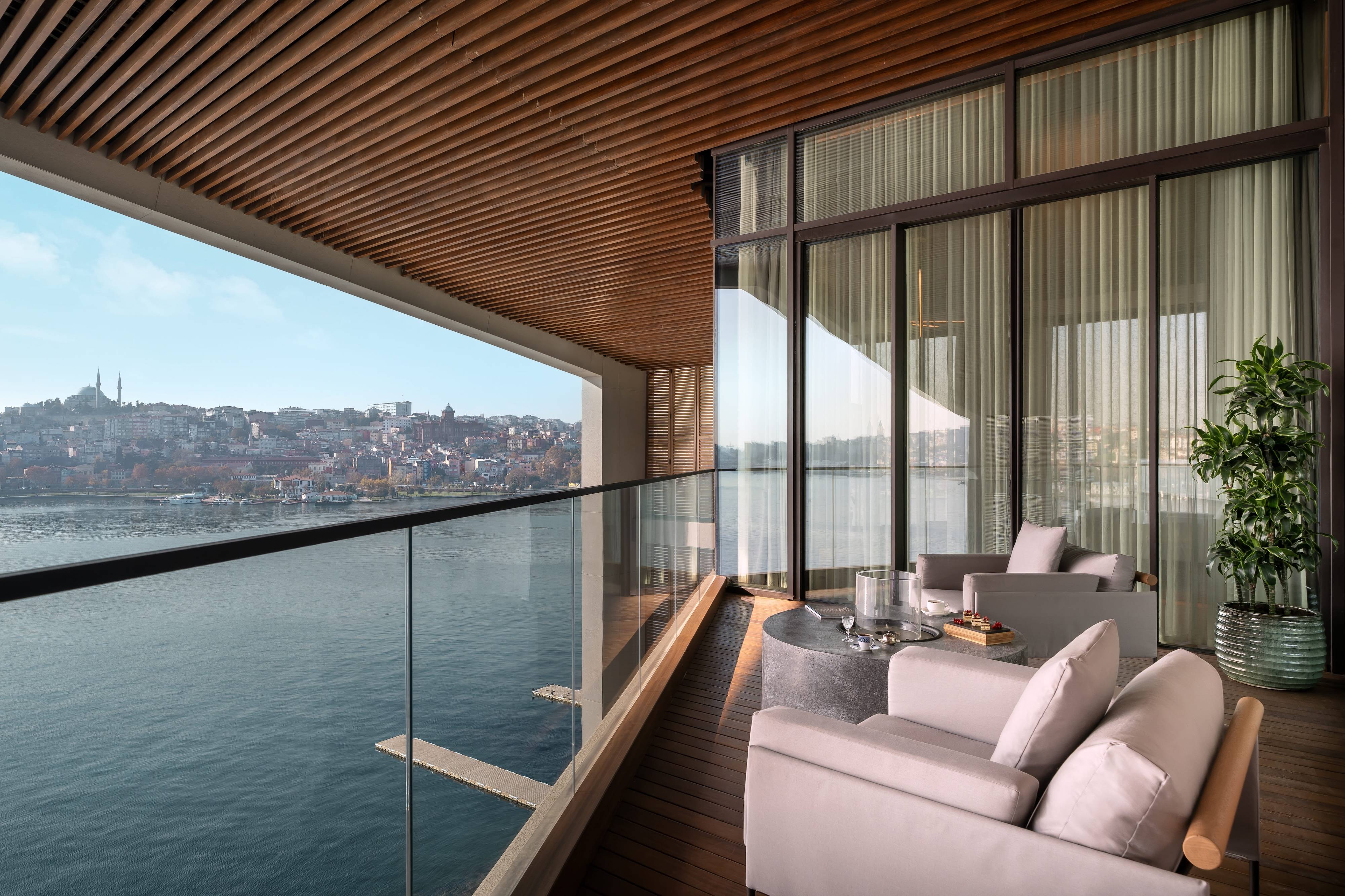 Nowoczesny apartament Dublex Terrace Suite z 1 sypialnią i widokiem na morze w hotelu Rixos Tersane Istanbul dysponuje balkonem ze szklanymi balustradami, wyściełanymi krzesłami, małym stolikiem i roślinami doniczkowymi, z którego roztacza się widok na wodę i miasto.