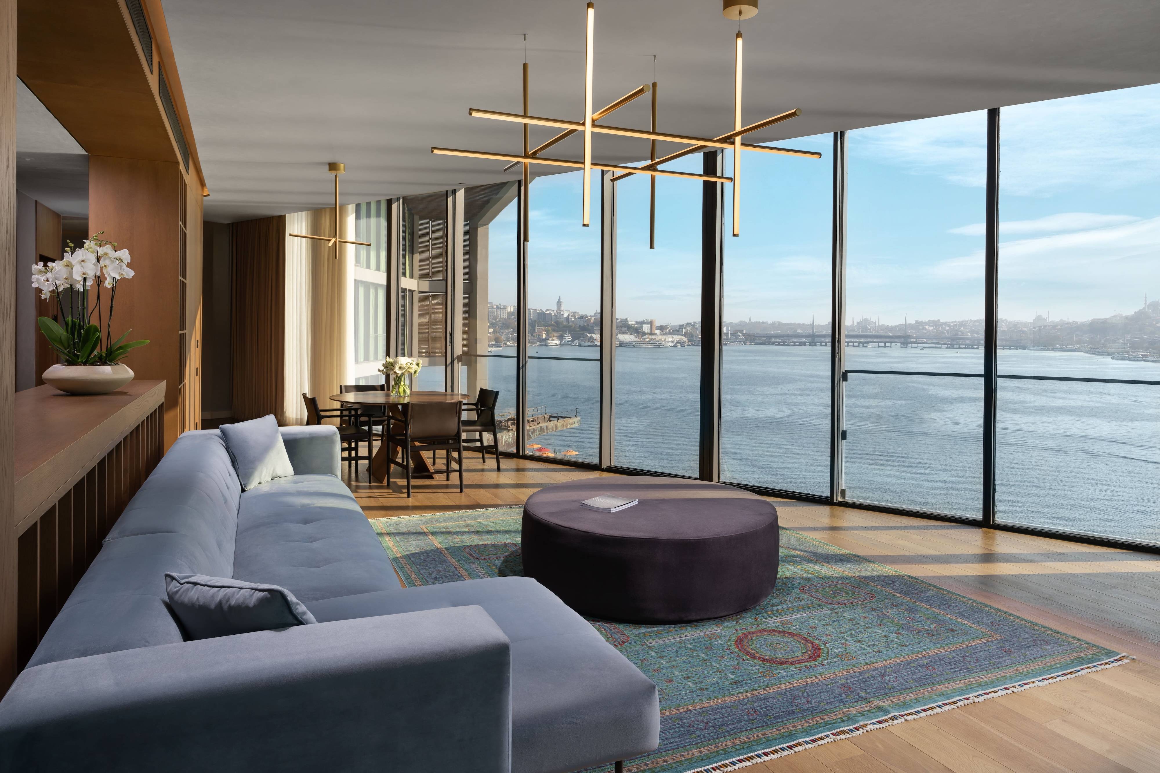 Nowoczesny salon w apartamencie Rixos Tersane Istanbul One Bedroom Dublex Terrace Suite with Sea View z dużymi oknami z widokiem na wodę, niebieską rozkładaną sofą, okrągłą otomaną, stołem jadalnym, złotymi żyrandolami i wzorzystym dywanem na drewnianej podłodze.