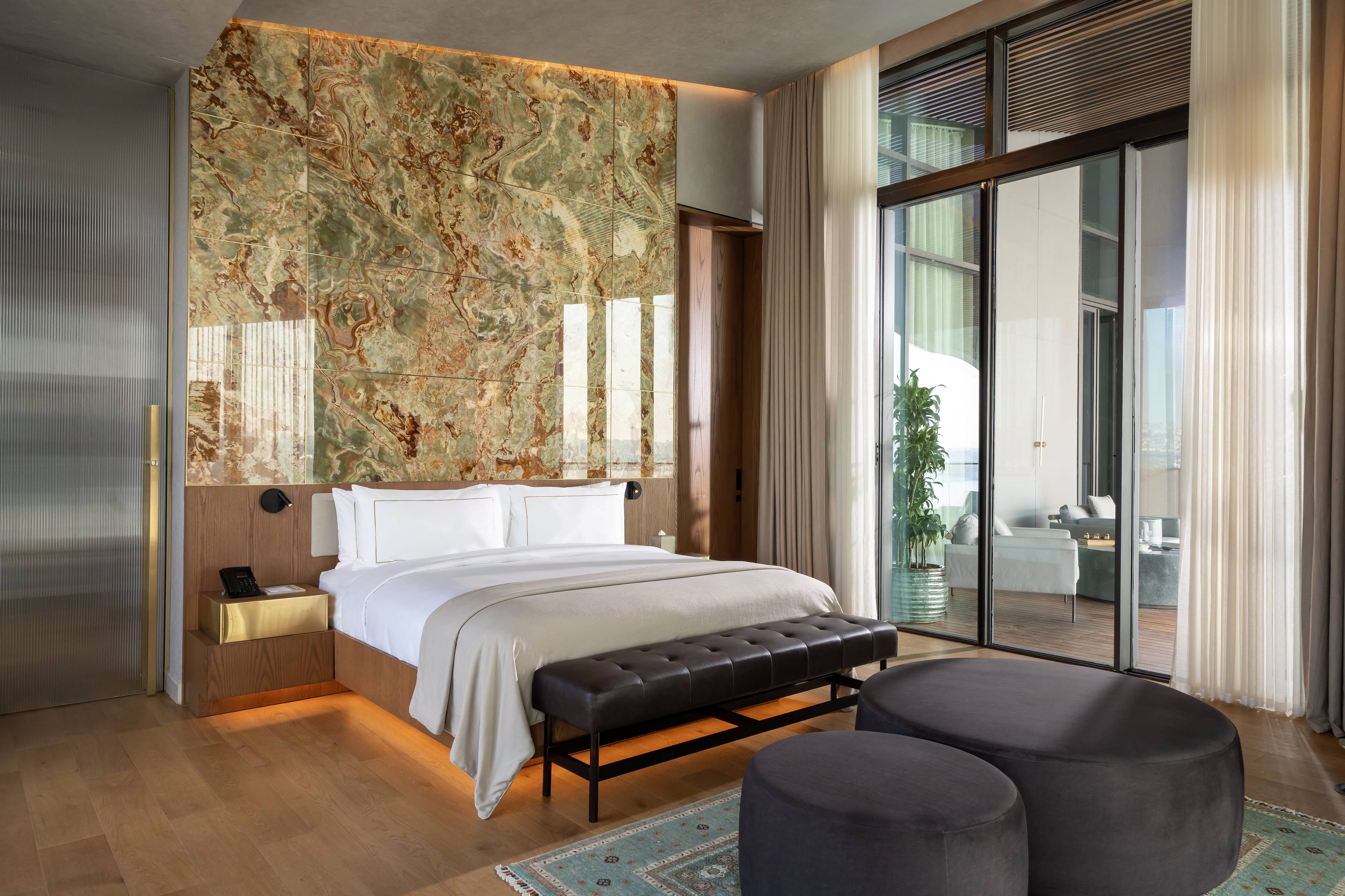 Nowoczesny apartament Rixos Tersane Istanbul One Bedroom Dublex Terrace Suite z widokiem na morze wyposażony jest w duże łóżko, kamienną ścianę akcentującą, okna sięgające od podłogi do sufitu wychodzące na balkon, neutralne kolory, czarną ławkę, okrągłe tafty i bujną zieloną roślinę na zewnątrz.