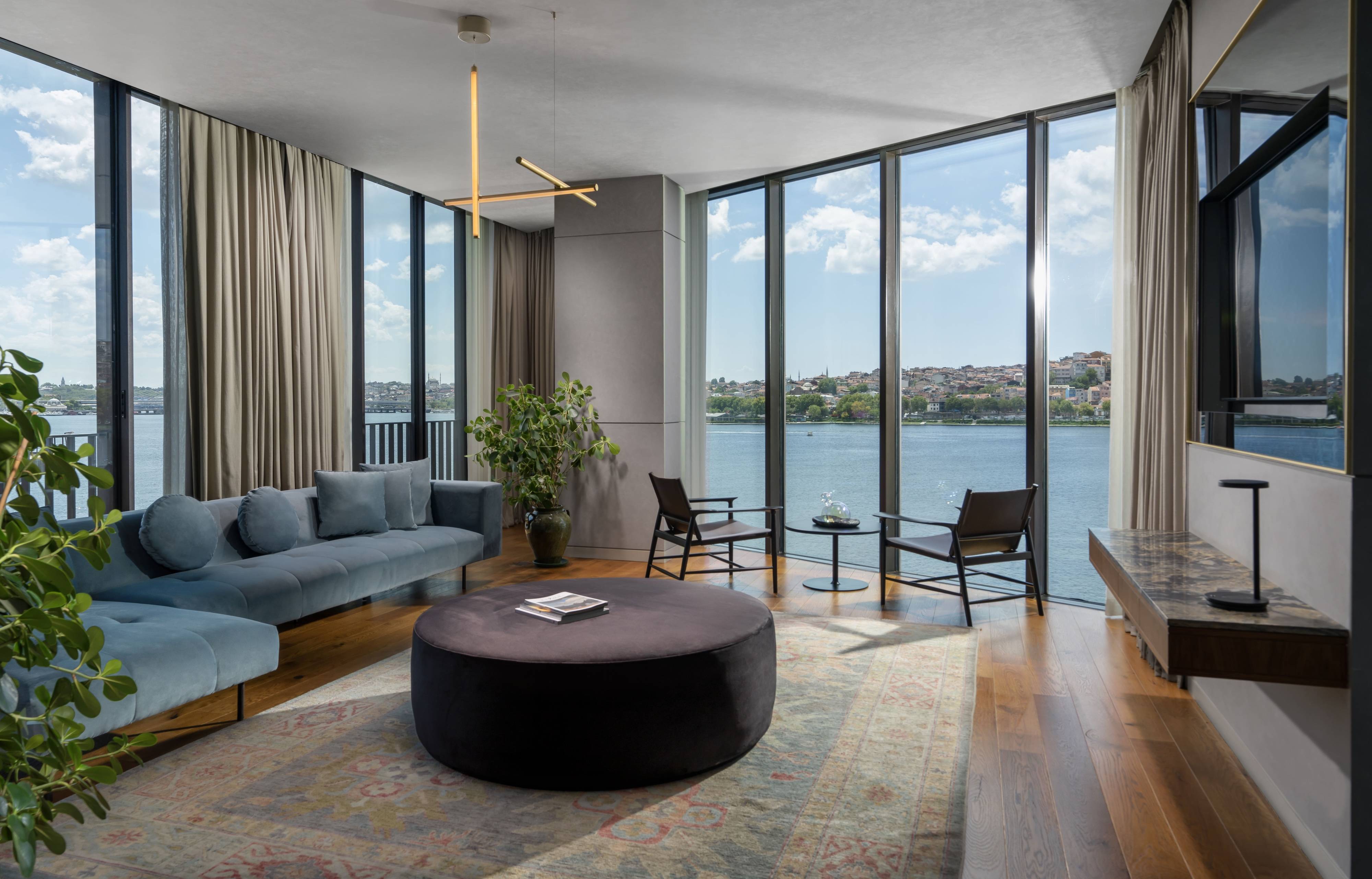 Modern living room in the One Bedroom Executive Terrace Suite Sea View at Rixos Tersane Istanbul features floor-to-ceiling windows, blue sectional sofa, round black ottoman, indoor plants, wall-mounted TV, and light wood floors in a bright, airy setting.