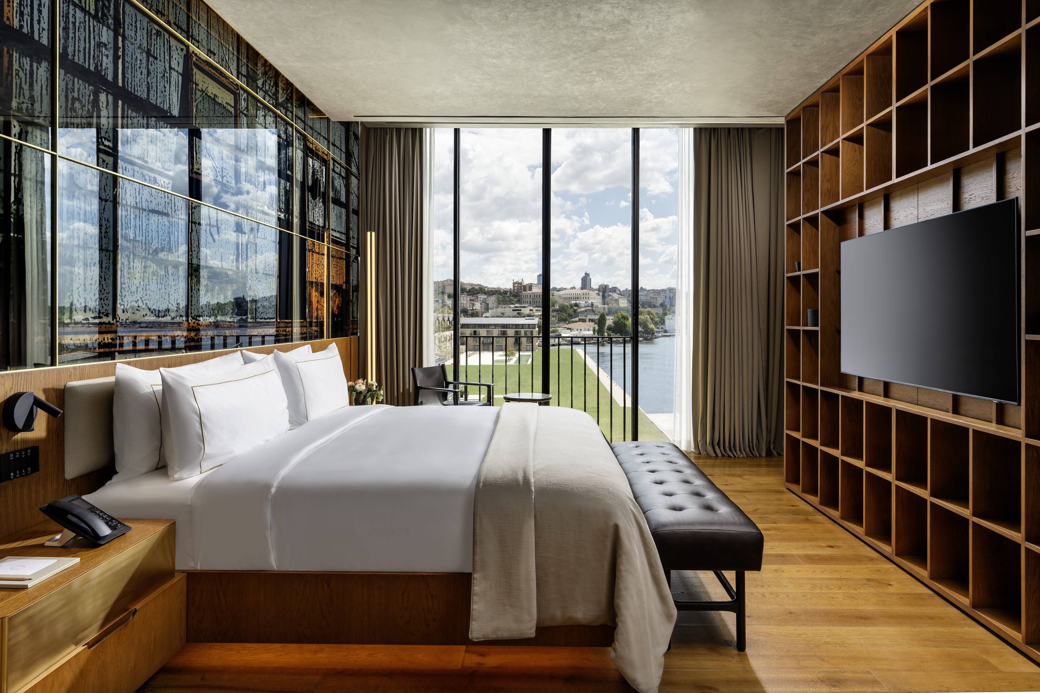 A modern One Bedroom Executive Terrace Suite Sea View at Rixos Tersane Istanbul features a large bed, wooden floors, wall-mounted TV, shelves, and floor-to-ceiling windows with river and cityscape views, filling the elegant space with natural light.