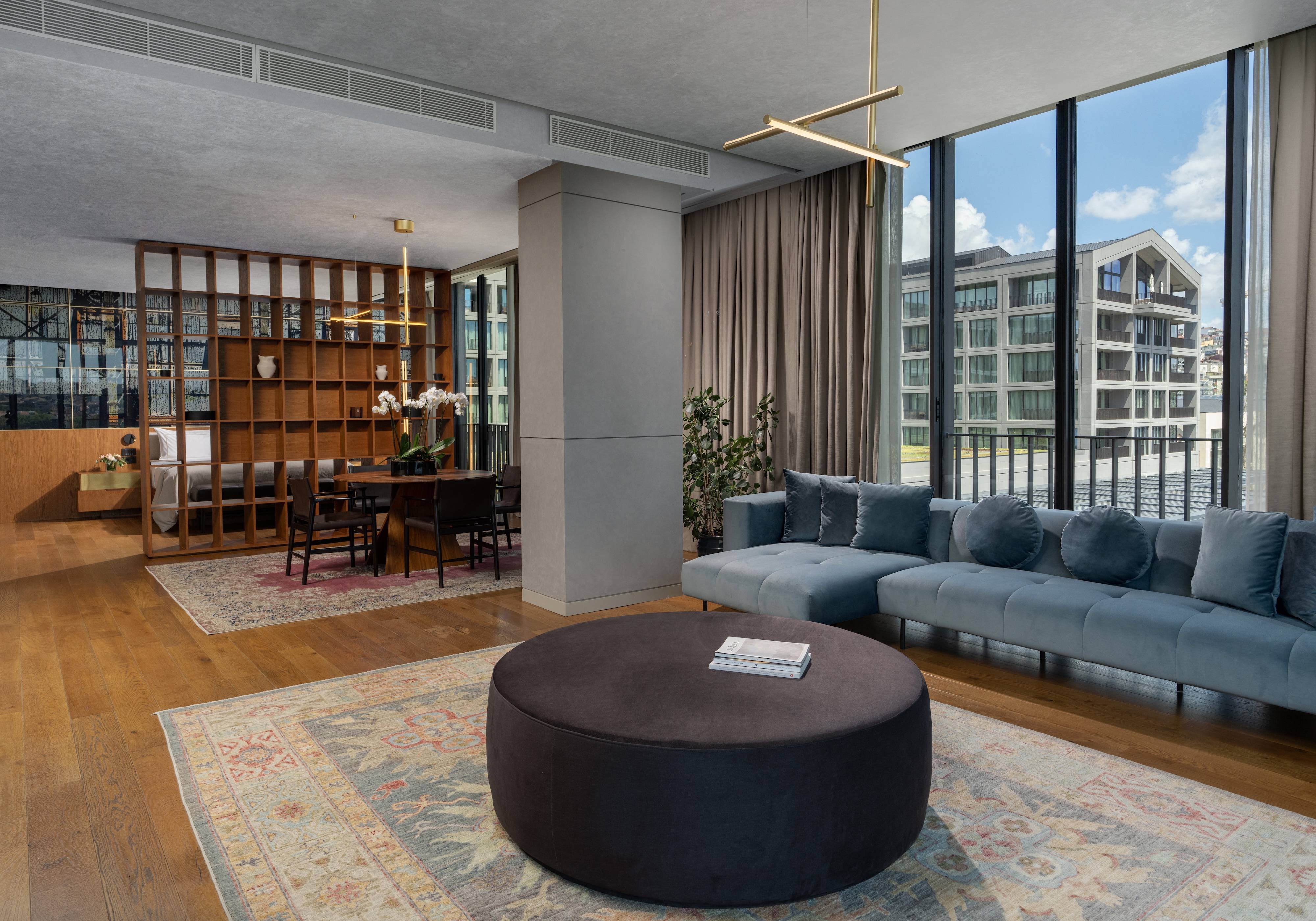 The One Bedroom Executive Terrace Suite Sea View at Rixos Tersane Istanbul features a spacious living area, blue sofa, round ottoman, large rug, dining table, and a bedroom separated by a wooden partition with city views through floor-to-ceiling windows.