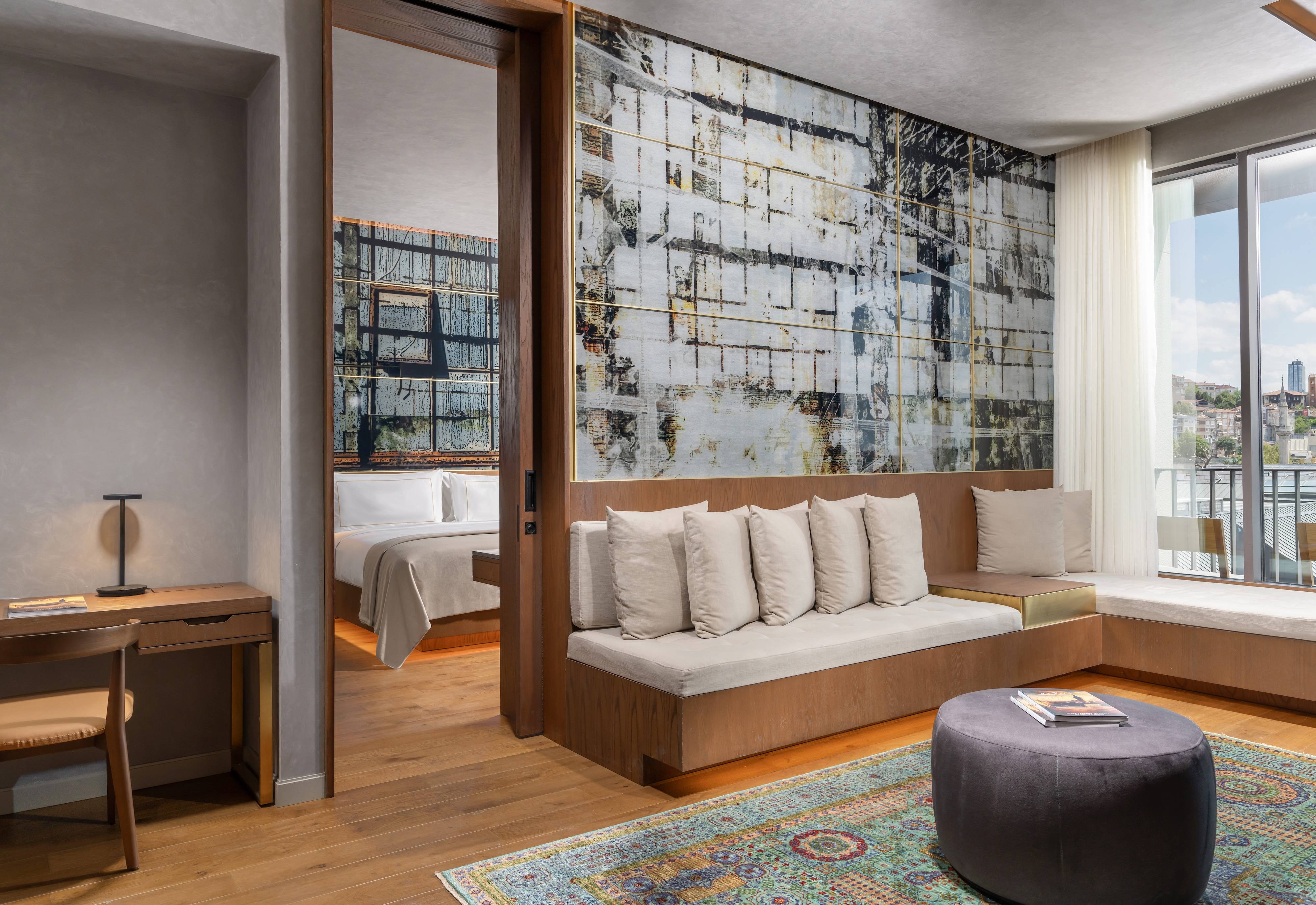 Nowoczesny apartament typu premium suite z 1 sypialnią i częściowym widokiem na morze oraz balkonem w hotelu Rixos Tersane Istanbul obejmuje dużą abstrakcyjną grafikę, wbudowaną kanapę z białymi poduszkami, okrągły otomana, kolorowy dywan, biurko oraz oddzielną sypialnię z otwartymi przesuwanymi drzwiami.