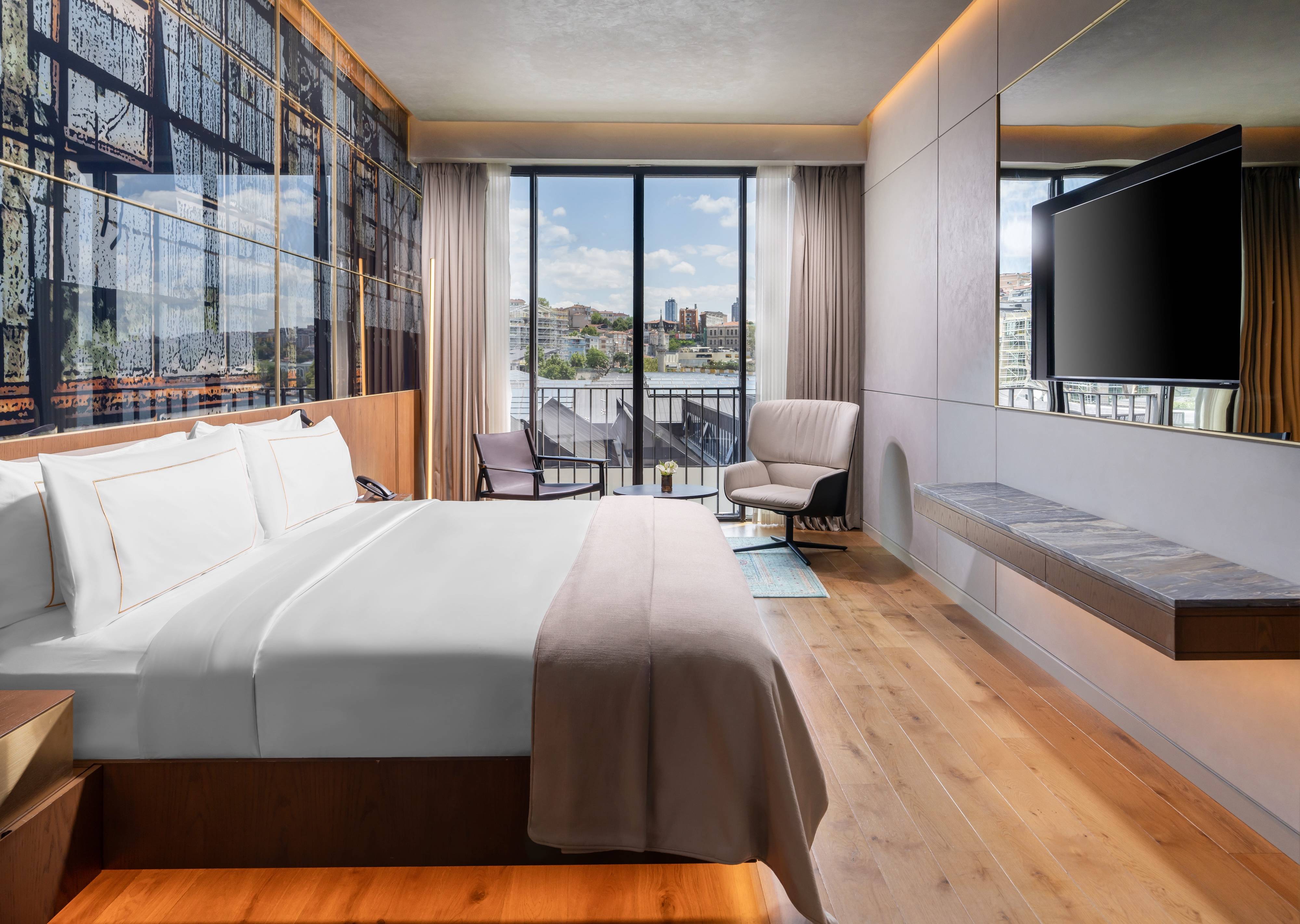 Modern Rixos Tersane Istanbul hotel room with a large bed, wall-mounted TV, wooden floors, lounge chair, and floor-to-ceiling windows offering a city view under a blue sky—One Bedroom Premium Suite Partial Sea View with Balcony.