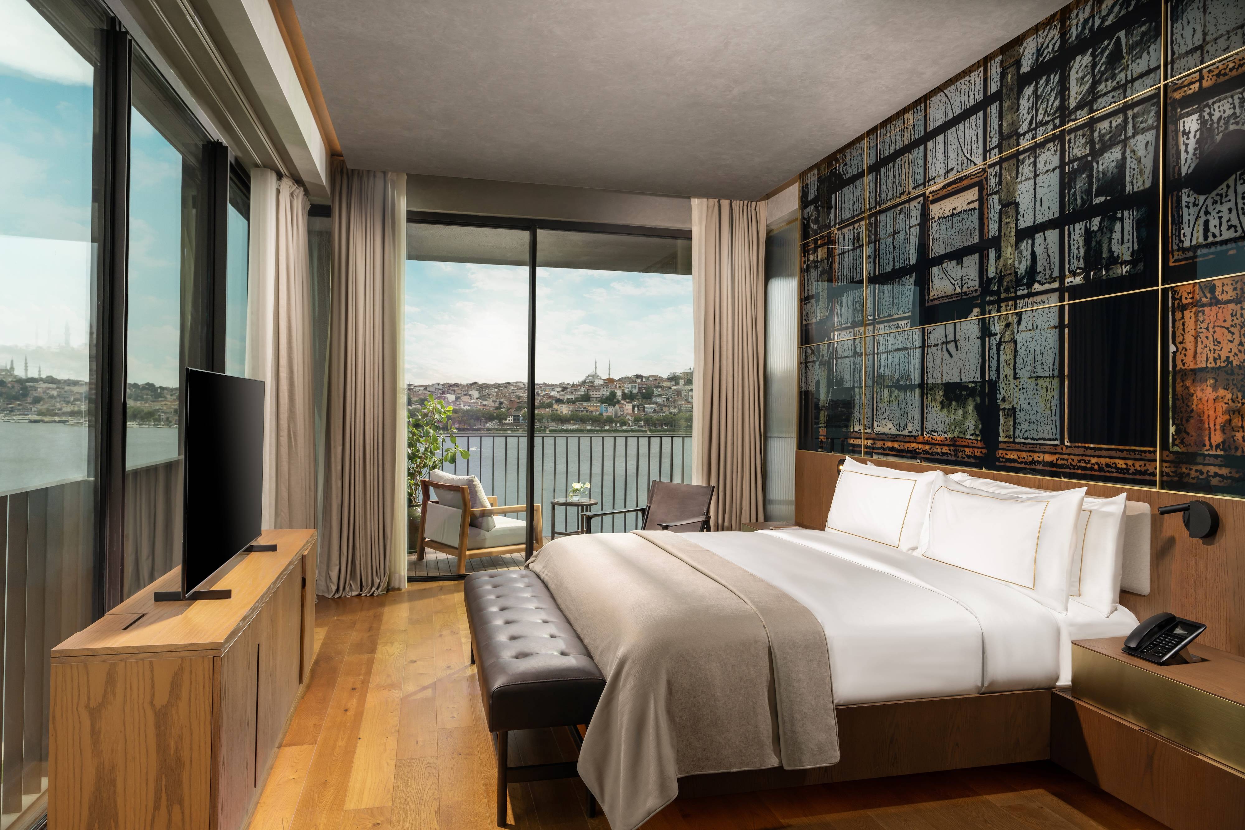 Apartament z 1 sypialnią Premium Terrace Suite Sea View w hotelu Rixos Tersane Istanbul to nowoczesny pokój z dużym łóżkiem, minimalistycznymi drewnianymi meblami, oknami sięgającymi od podłogi do sufitu, balkonem z widokiem na miasto i wodę oraz artystycznym muralem skąpanym w naturalnym świetle.