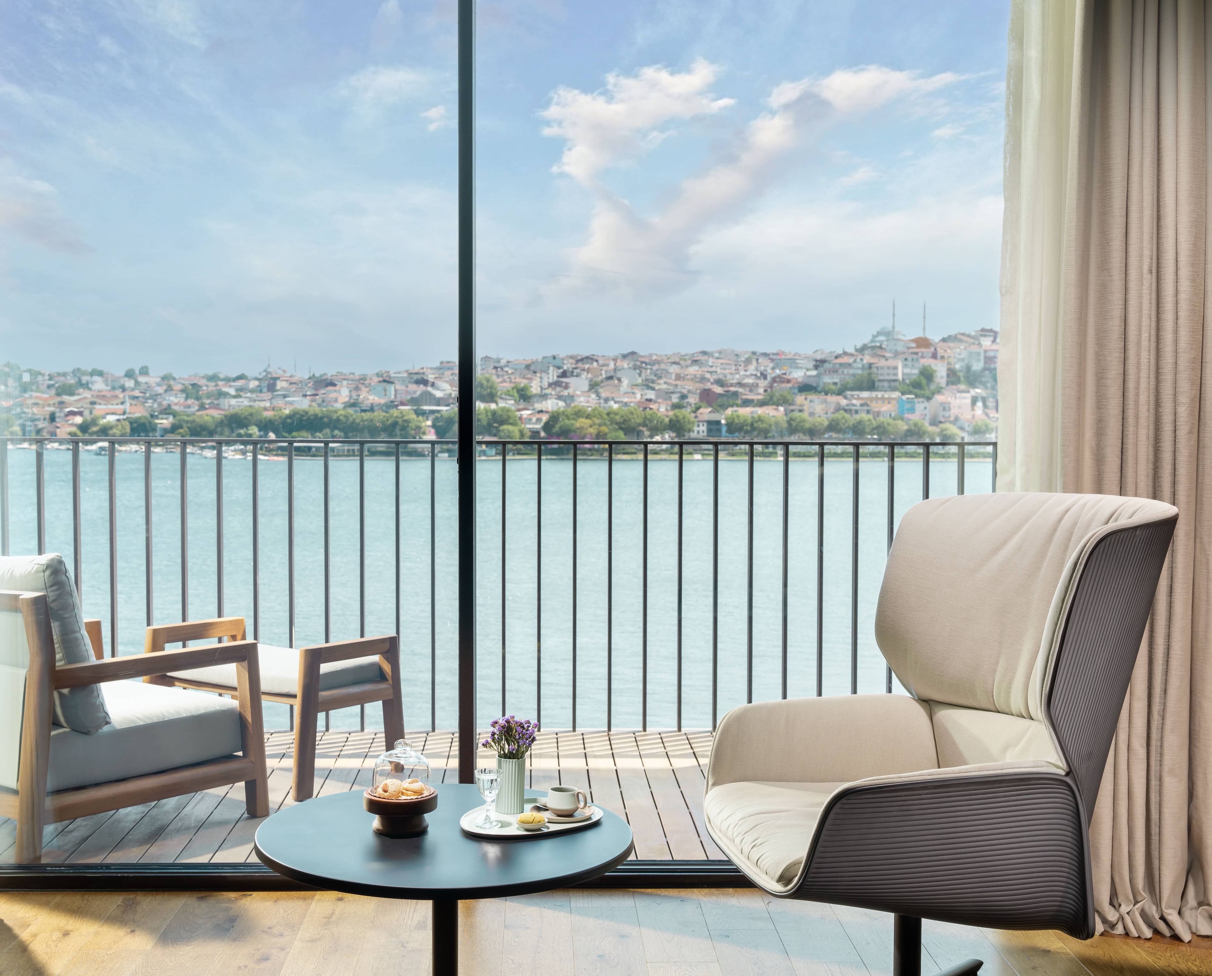 Modern lounge area in the One Bedroom Premium Terrace Suite Sea View at Rixos Tersane Istanbul, with armchairs and a round table set for tea, overlooking a scenic river, cityscape, and blue sky through large glass windows.