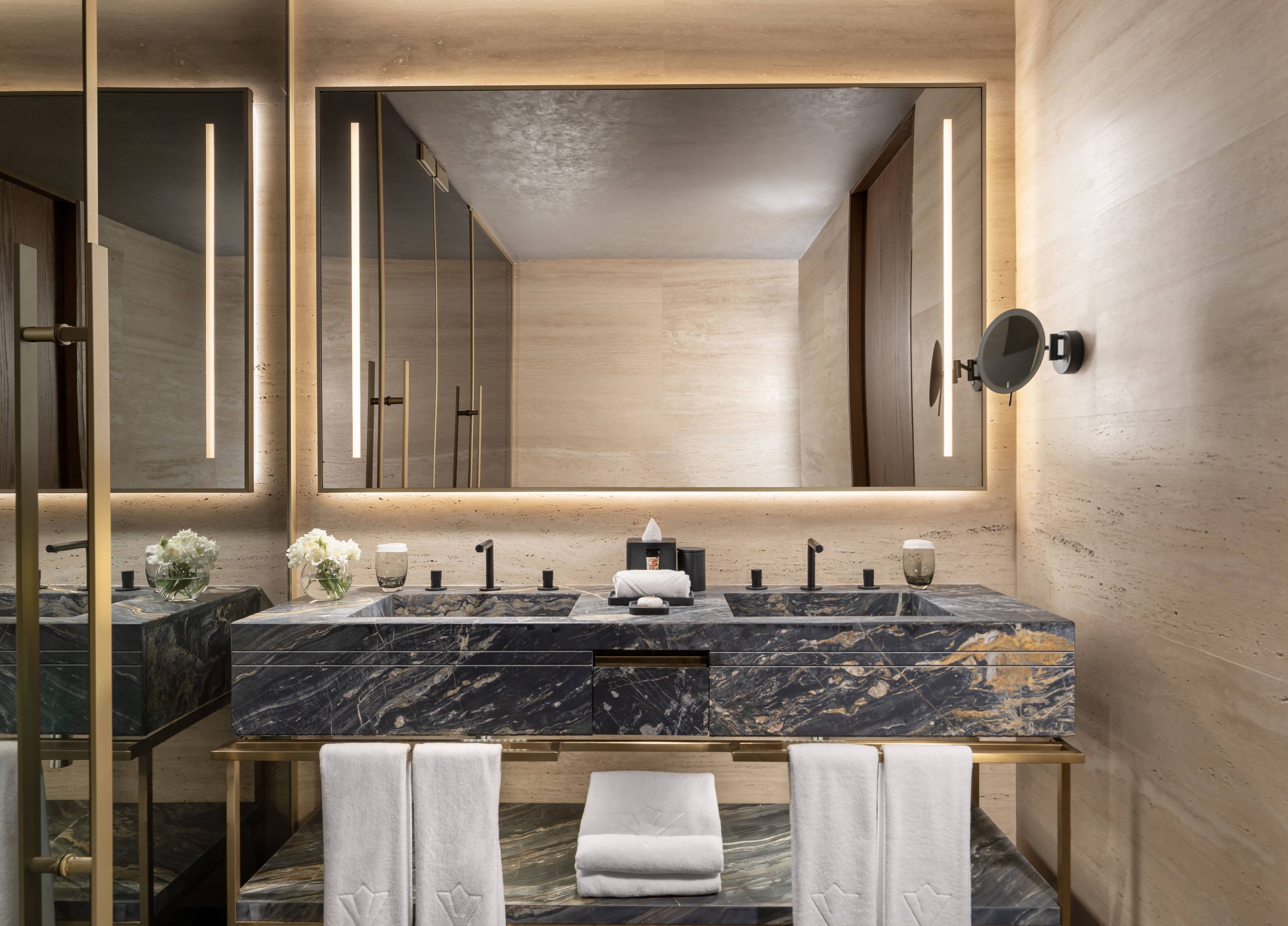 Modern bathroom in the One Bedroom Premium Terrace Suite Sea View at Rixos Tersane Istanbul, featuring a double marble sink, large illuminated mirror, fresh flowers, and beige stone walls for a sleek, luxurious atmosphere.