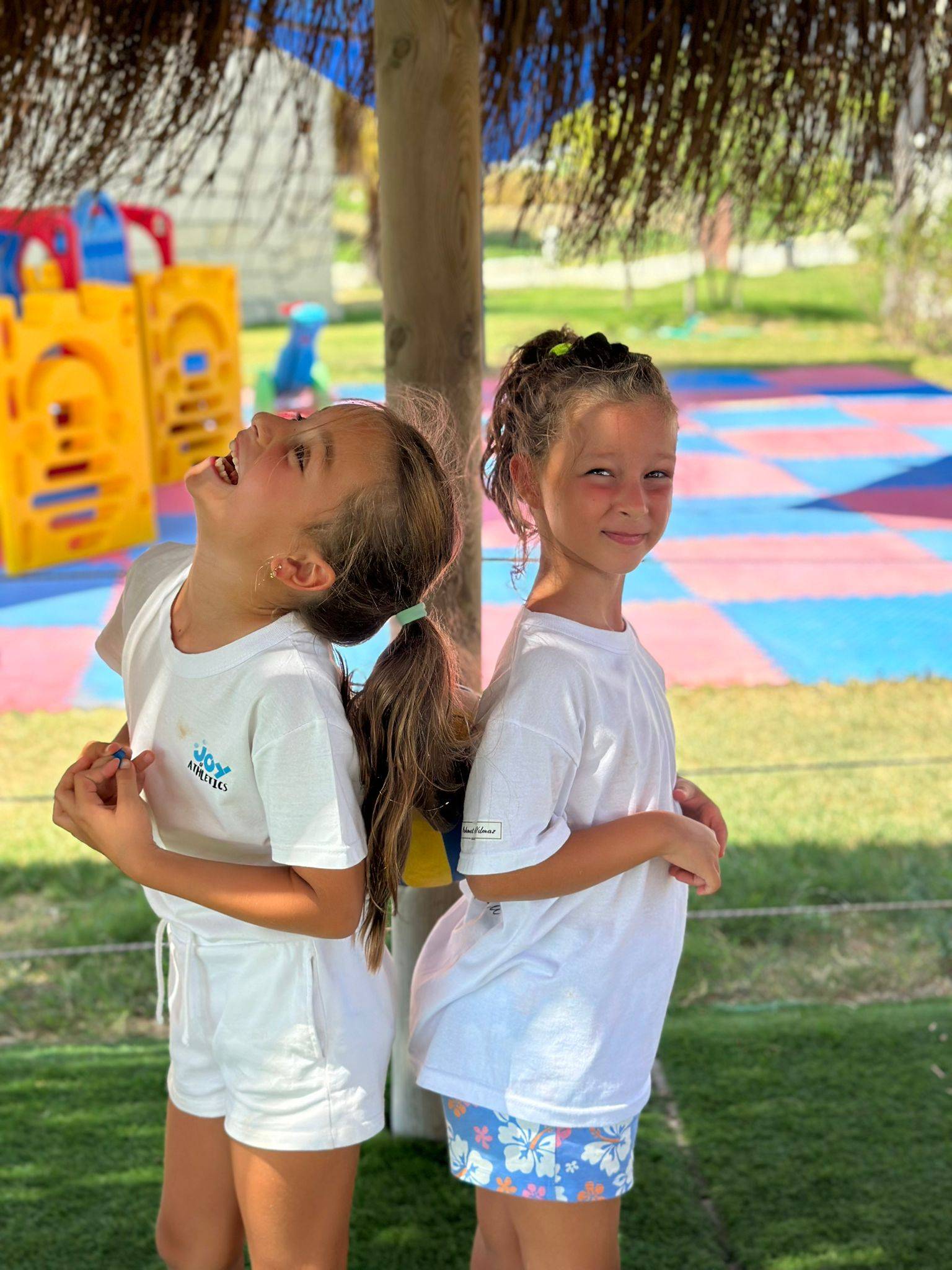 Dwie młode dziewczyny stoją plecami do siebie, uśmiechając się i śmiejąc pod zacienionym obszarem na placu zabaw, promieniując radością z lekkiej atletyki. Obie mają na sobie białe koszulki i szorty, a w tle widać kolorowe wyposażenie placu zabaw i matę w kratkę.
