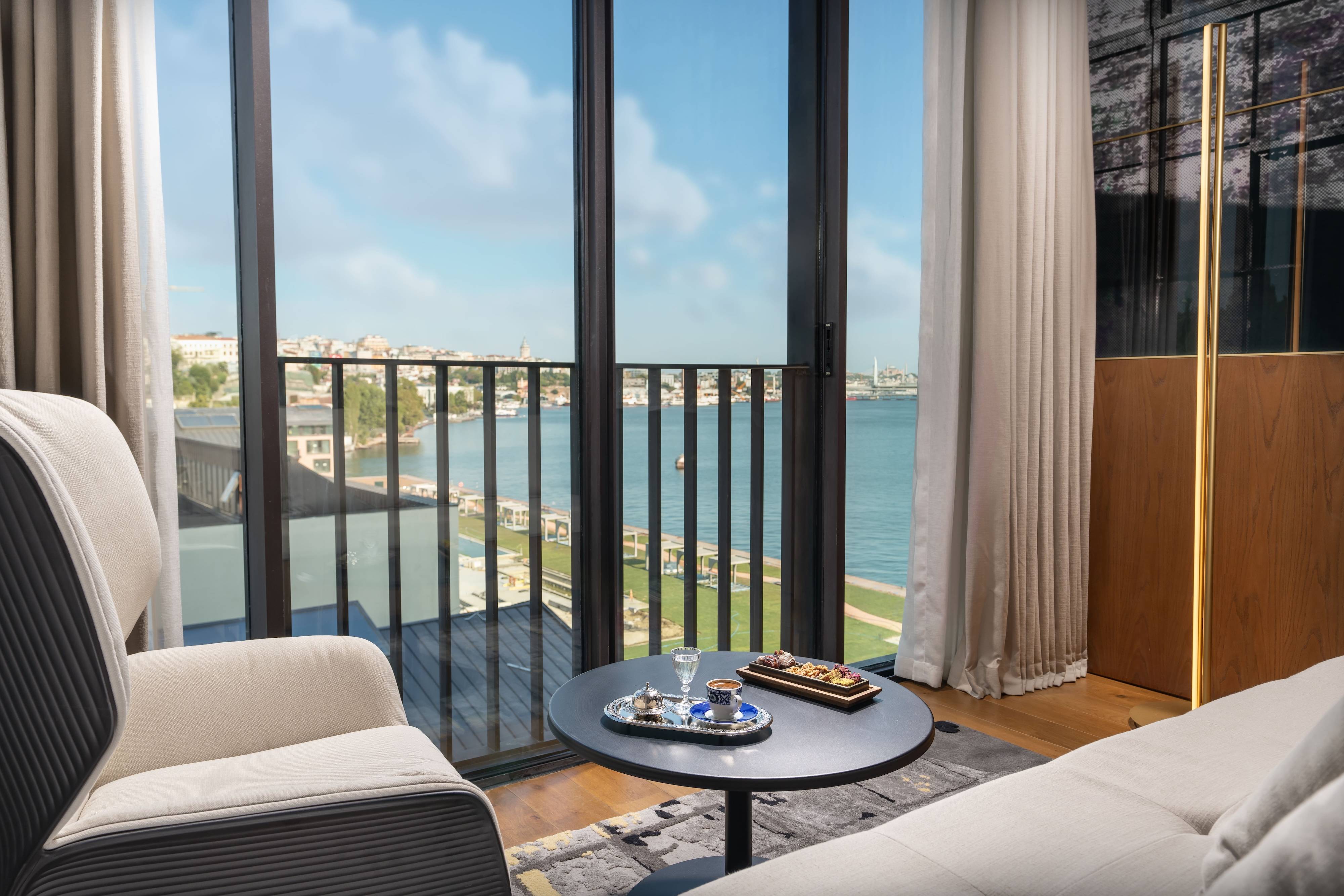 A cozy living room at Rixos Tersane Istanbul features armchairs and a table set with coffee and treats, overlooking a balcony with a scenic view of the river, city buildings, and a clear blue sky.