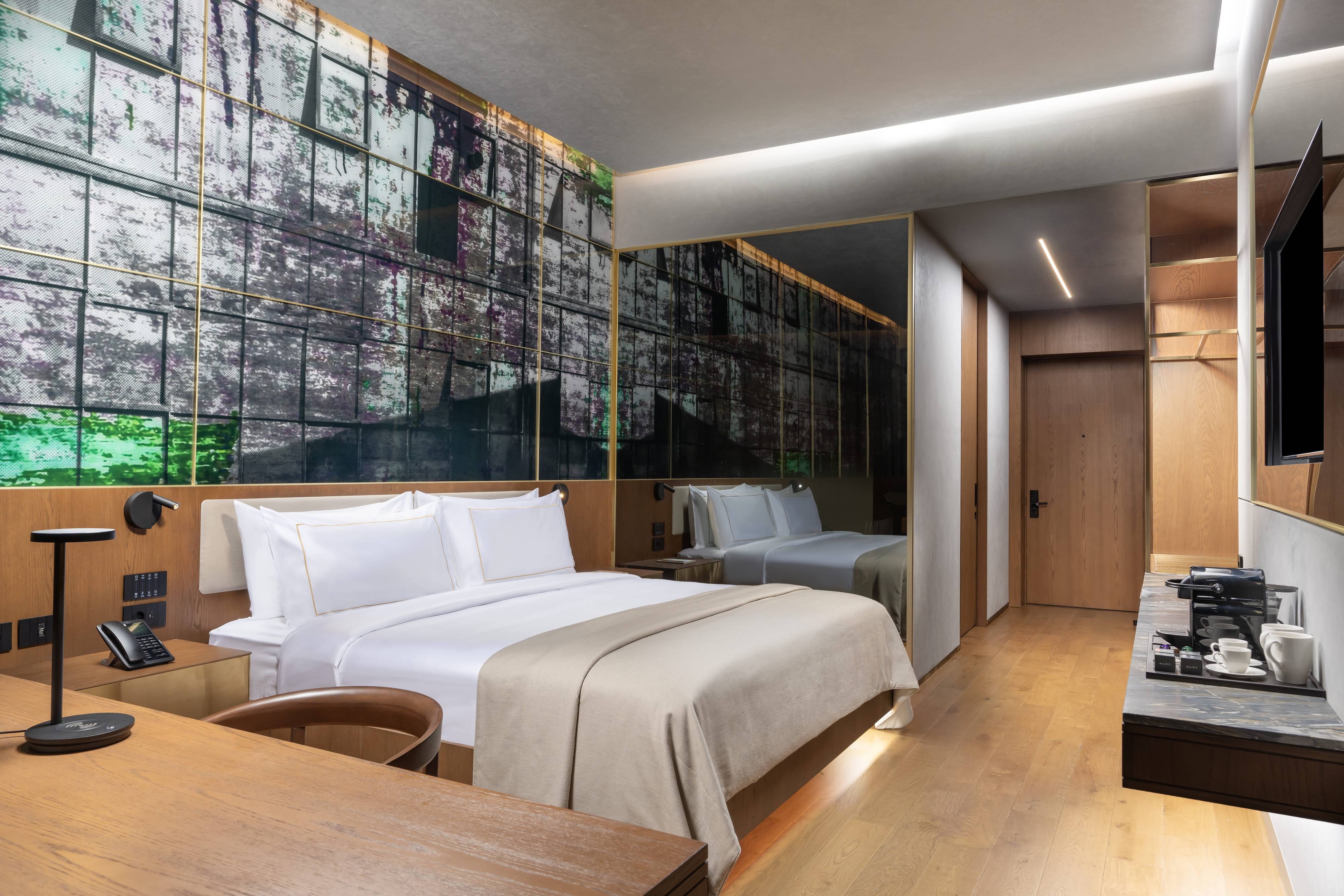 Nowoczesny pokój w hotelu Rixos Tersane z dużym łóżkiem z białą pościelą, beżową narzutą, drewnianymi meblami, abstrakcyjną fototapetą, lustrzaną ścianą i stacją do kawy na marmurowym blacie. Pokój Premium z częściowym widokiem na morze typu king-size oferuje ciepłe oświetlenie.