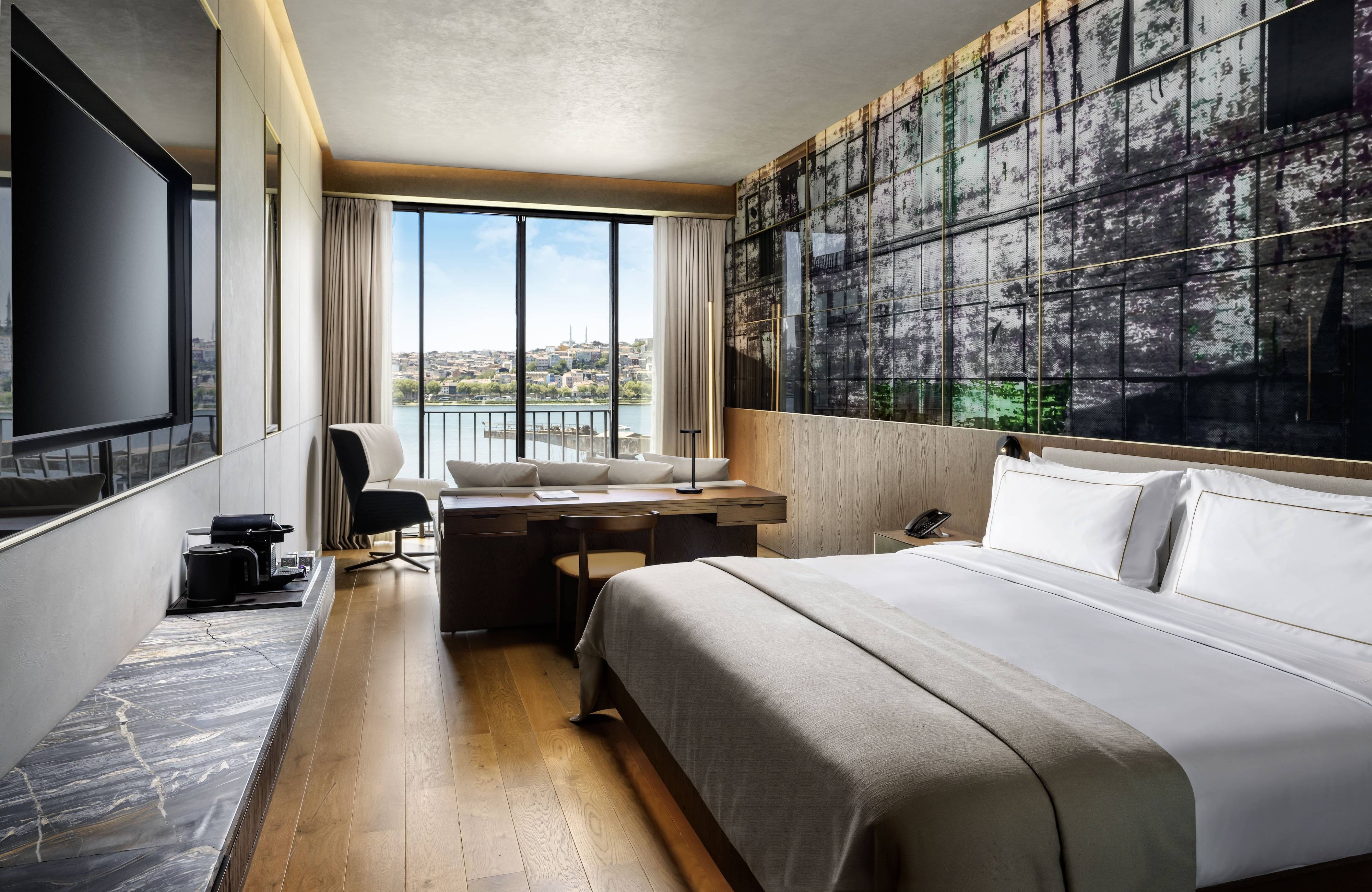 Experience the Premium Room Sea View King at Rixos Tersane Istanbul—a modern hotel room with a large bed, desk, flat-screen TV, coffee maker, and floor-to-ceiling windows framing a scenic river view in sleek, minimalist style.
