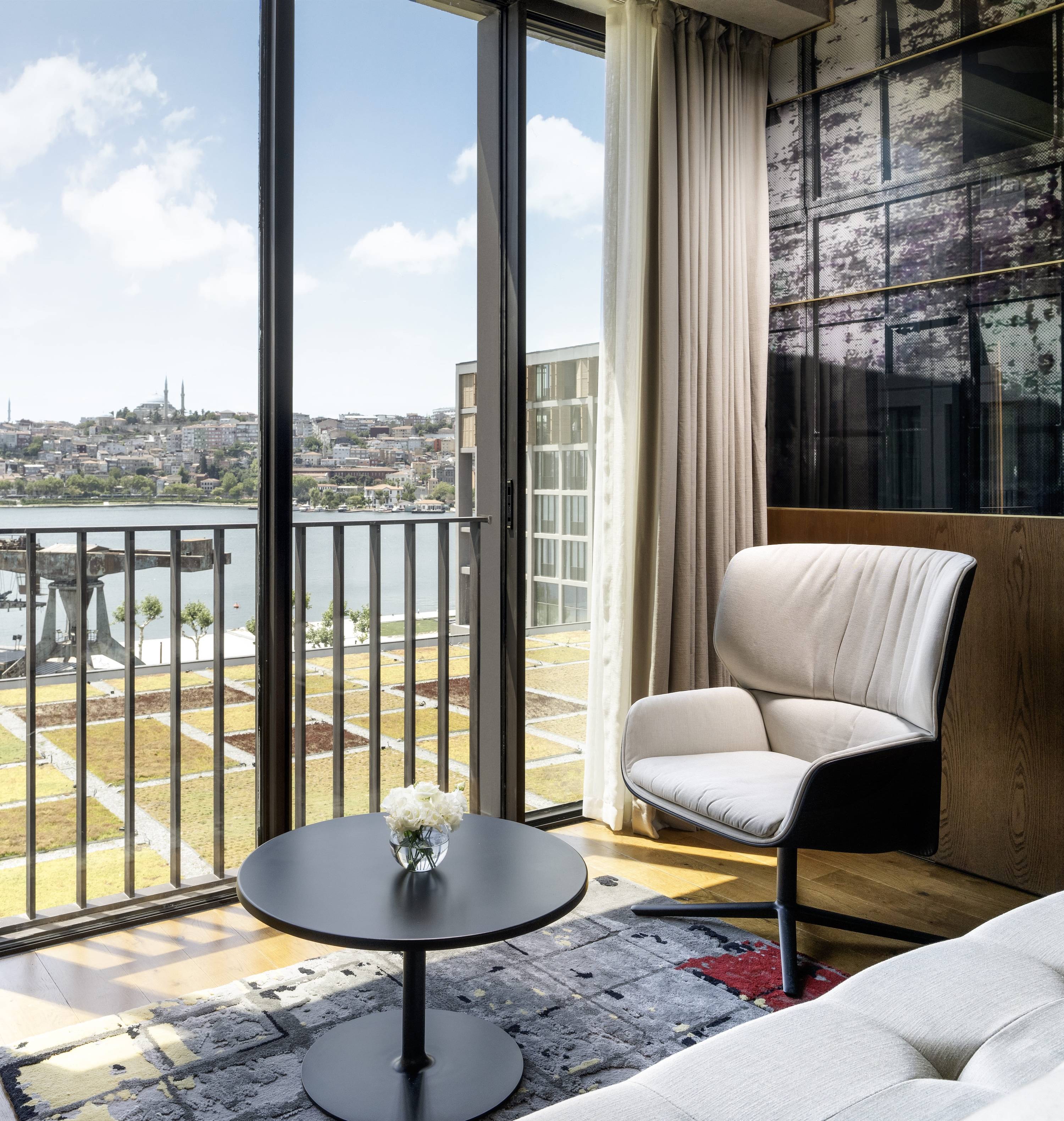 Nowoczesny salon w hotelu Rixos Tersane Istanbul Premium Room Sea View King wyposażony jest w biały fotel, okrągły czarny stół z kwiatami, wzorzyste ściany, kolorowy dywan i duże okna, przez które można podziwiać widoki na miasto i nabrzeże.