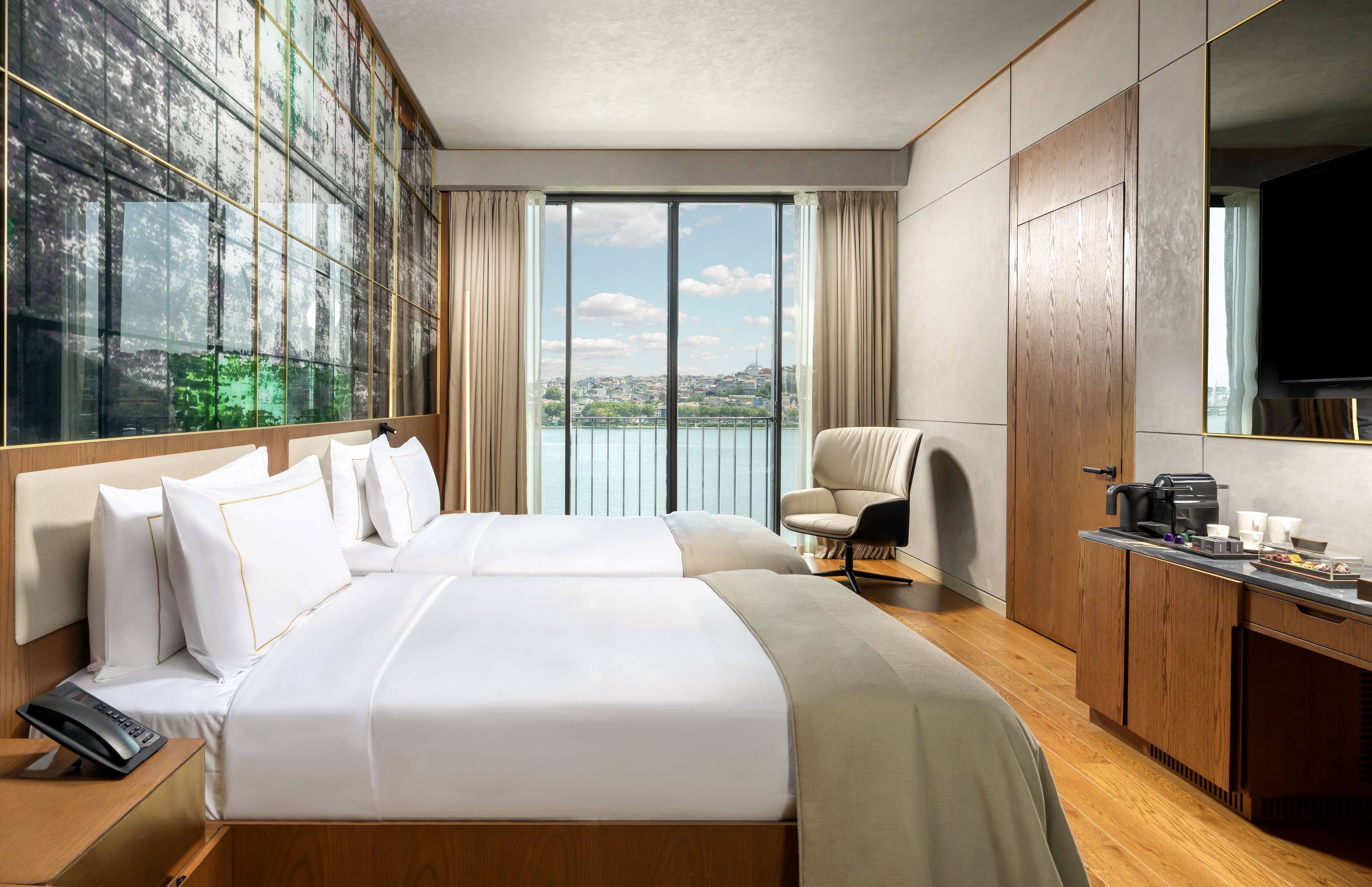 Nowoczesny pokój Premium z widokiem na morze Twin w hotelu Rixos Tersane Istanbul wyposażony jest w dwa schludnie pościelone łóżka, wygodny fotel, stację do kawy na drewnianej szafce, podłogi z jasnego drewna i ciepły wystrój - a wszystko to z pięknym widokiem na miasto i wodę przez duże okno.