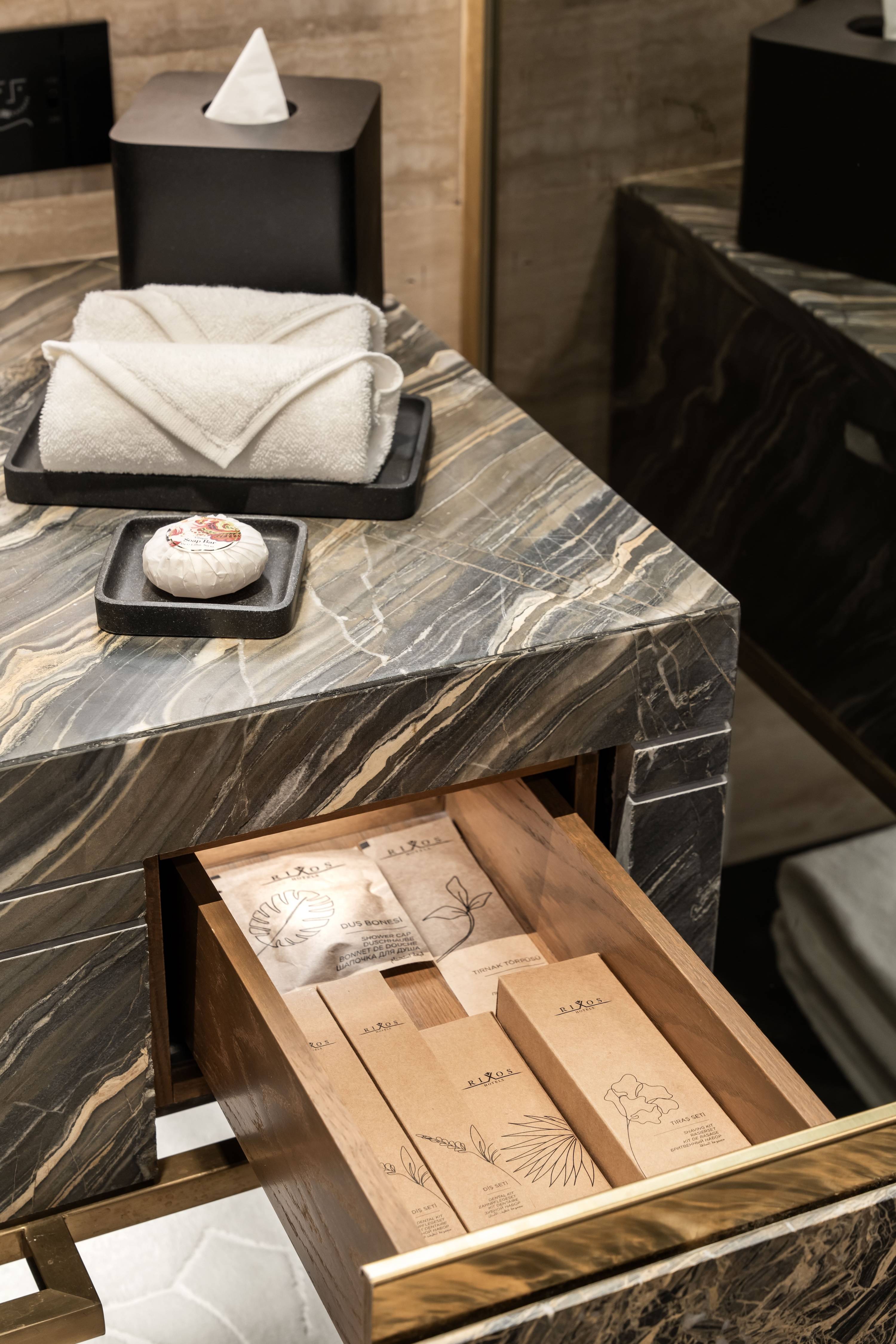 A marble bathroom countertop in the Premium Room Sea View Twin at Rixos Tersane Istanbul features a folded towel, a small tray with wrapped soap, and a tissue box. An open drawer reveals neatly arranged toiletries in brown paper packaging.