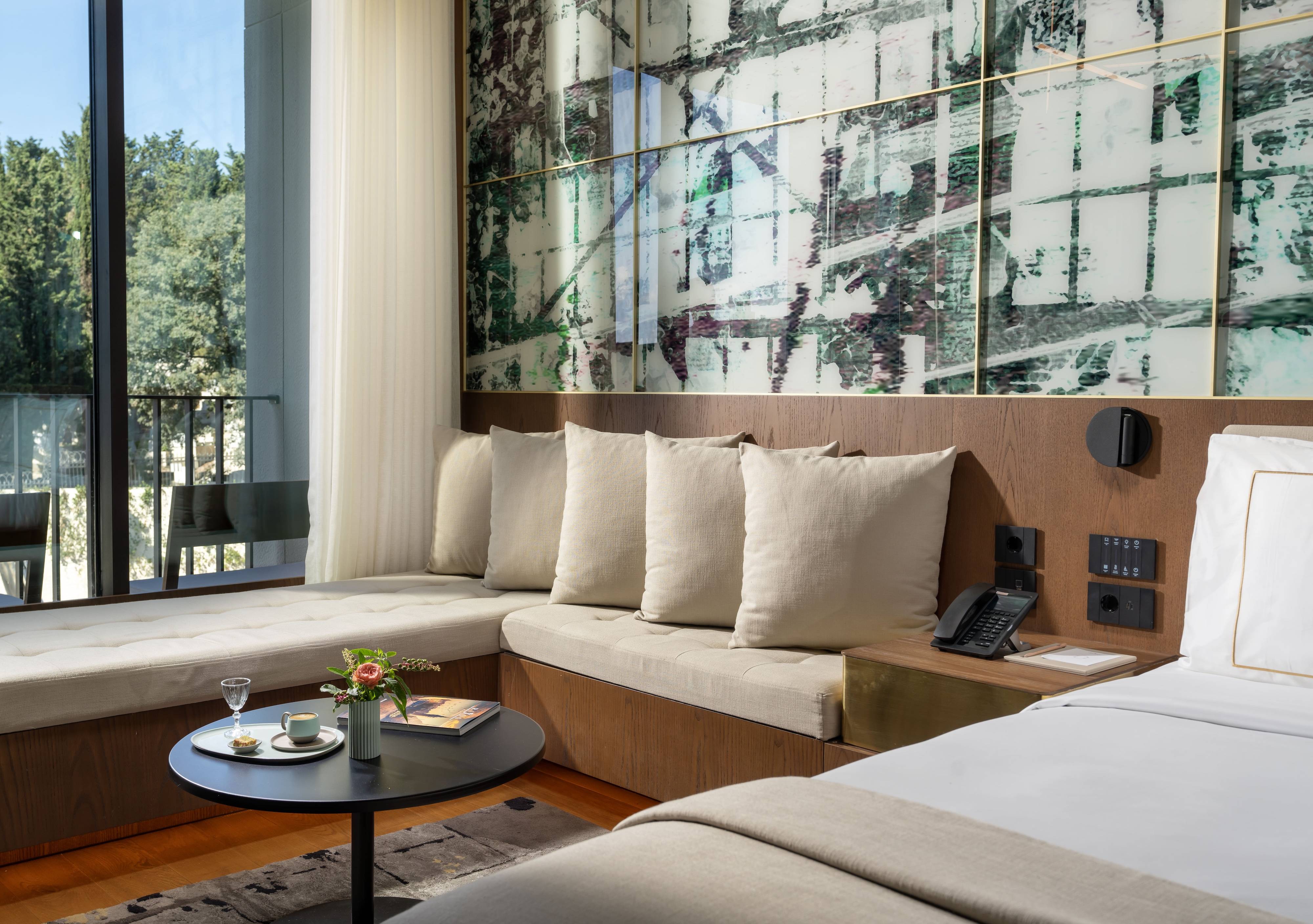 A modern Rixos Tersane Istanbul hotel room featuring a bed, cushioned corner seating, a round table set with coffee and pastries, a wall-mounted phone, and large windows overlooking trees and a balcony. Premium Room Twin