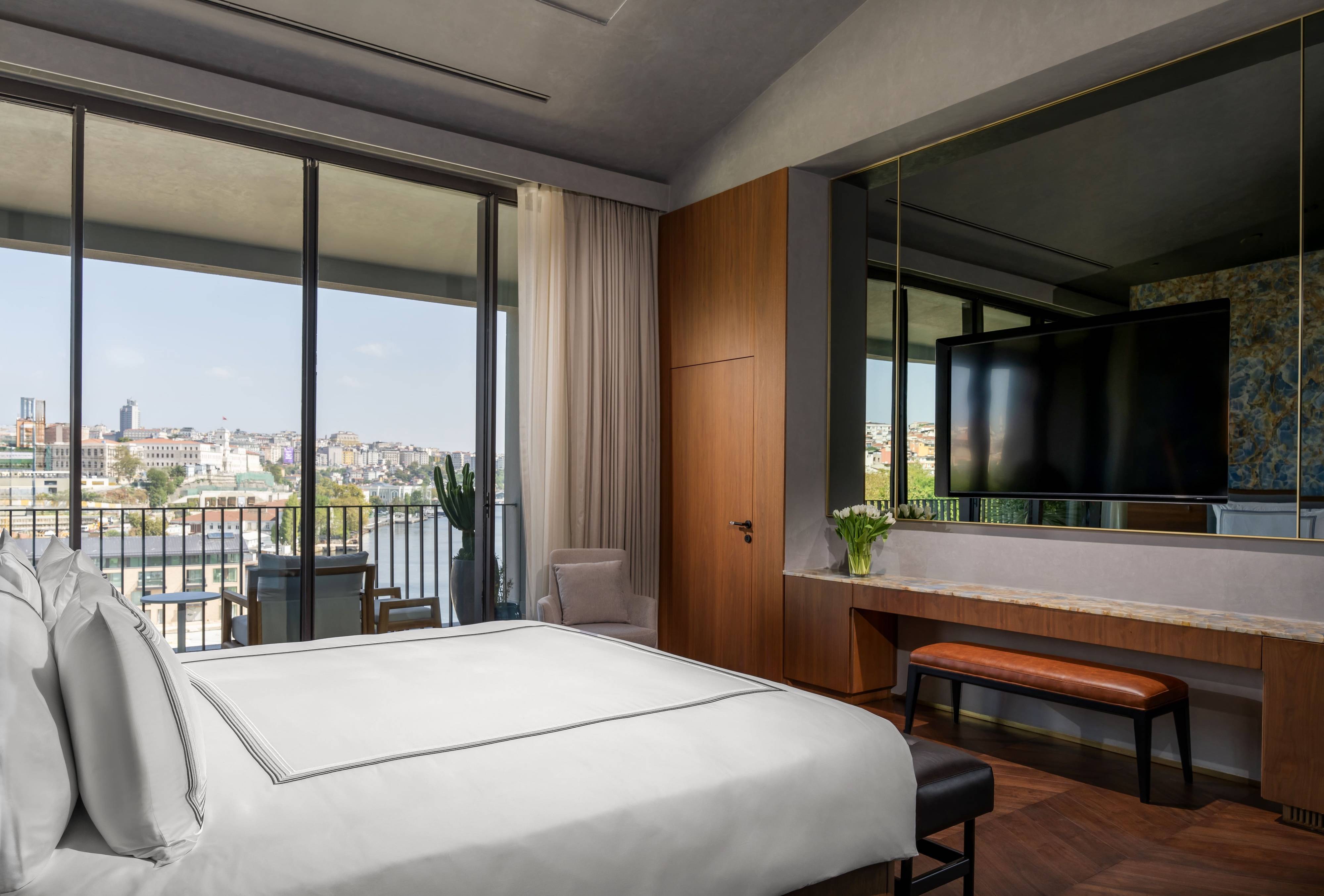 Experience the elegance of the Presidential Suite at Rixos Tersane Istanbul—this modern hotel room features a neatly made bed, large wall-mounted TV, and floor-to-ceiling windows offering a stunning city view and balcony access.