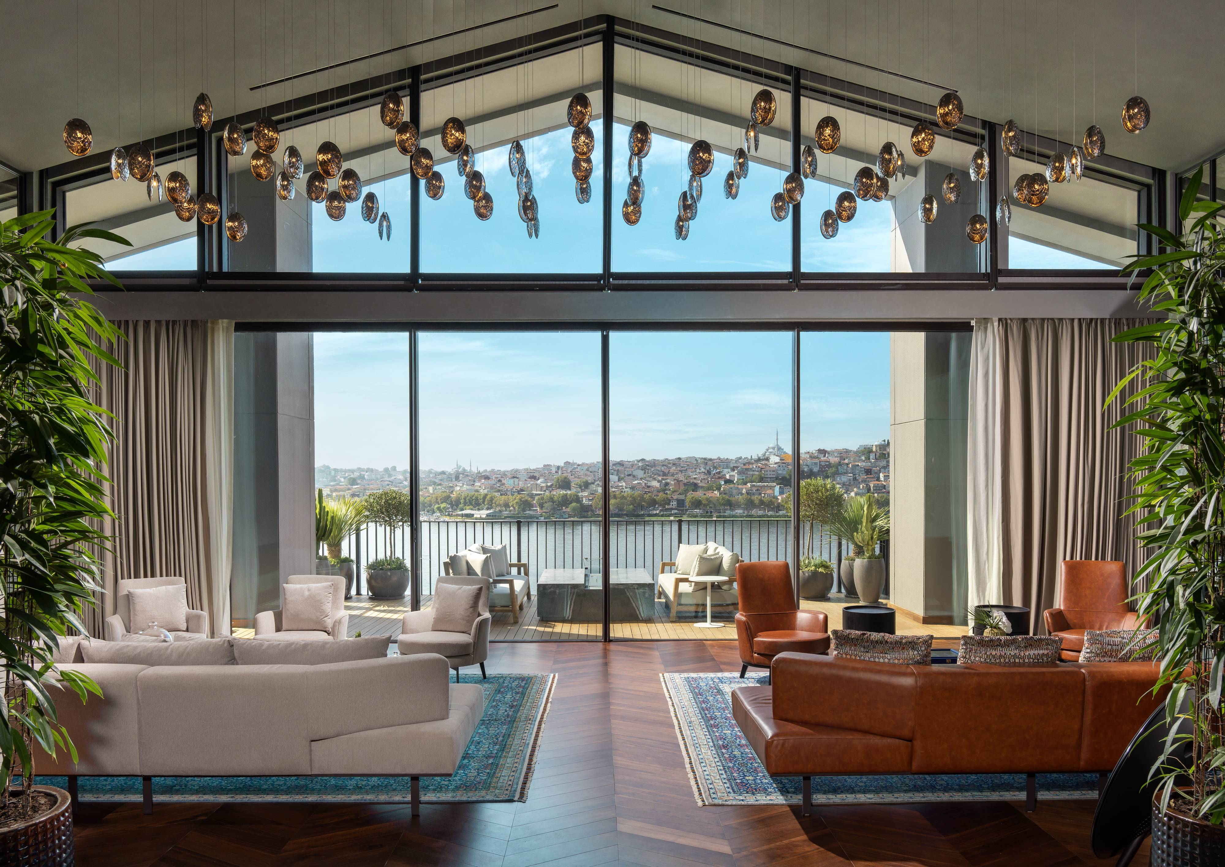 Nowoczesny salon w apartamencie prezydenckim Rixos Tersane Istanbul z dużymi oknami z widokiem na miasto i rzekę, beżowymi sofami, brązowymi skórzanymi fotelami, eleganckimi dywanami, bujnymi zielonymi roślinami i efektowną oprawą sufitową z wiszącymi żarówkami.