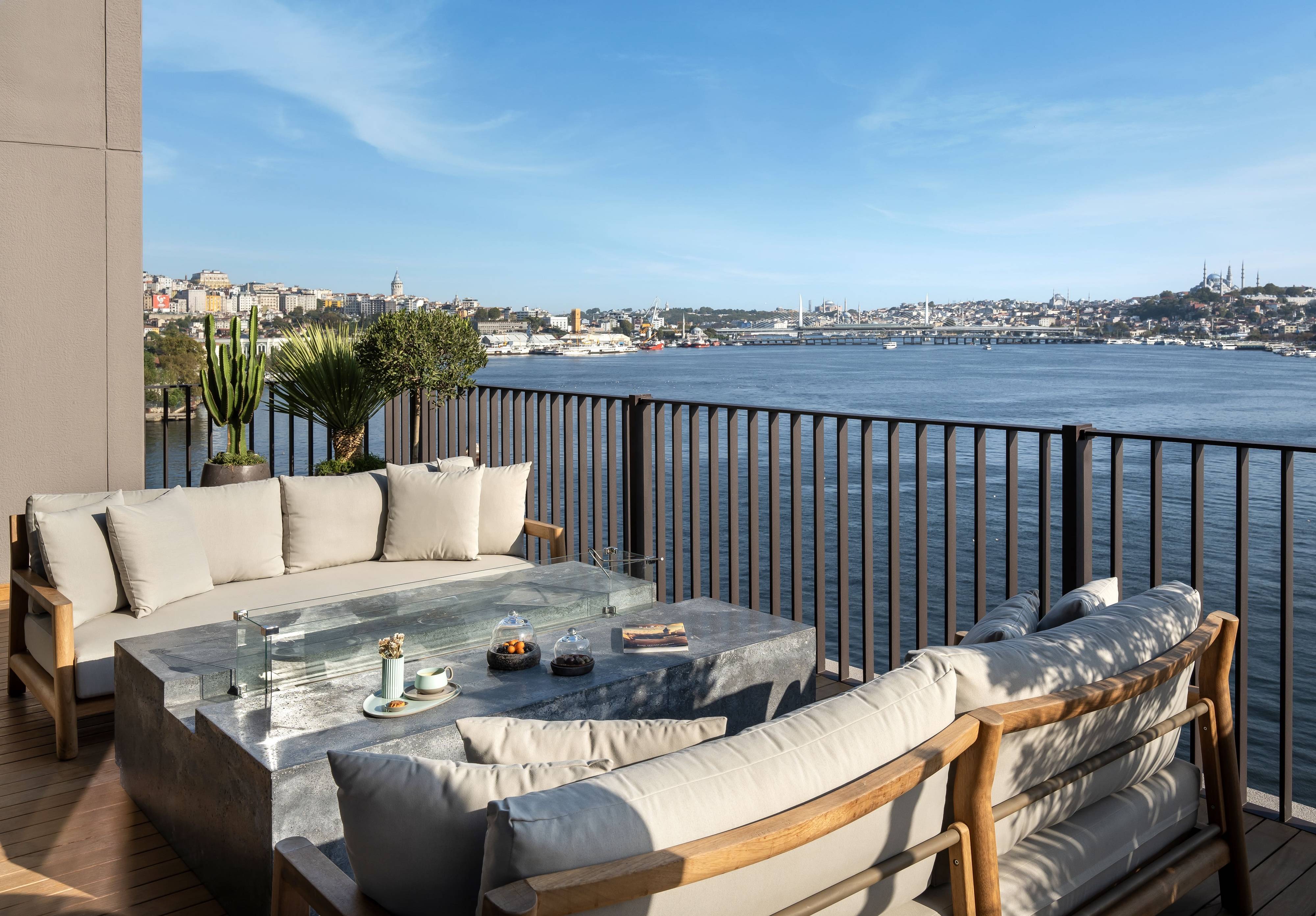 Na tarasie na dachu apartamentu prezydenckiego Rixos Tersane Istanbul znajdują się wyściełane drewniane sofy, betonowy stolik kawowy i rośliny doniczkowe, z widokiem na dużą rzekę i panoramę miasta pod czystym, błękitnym niebem.