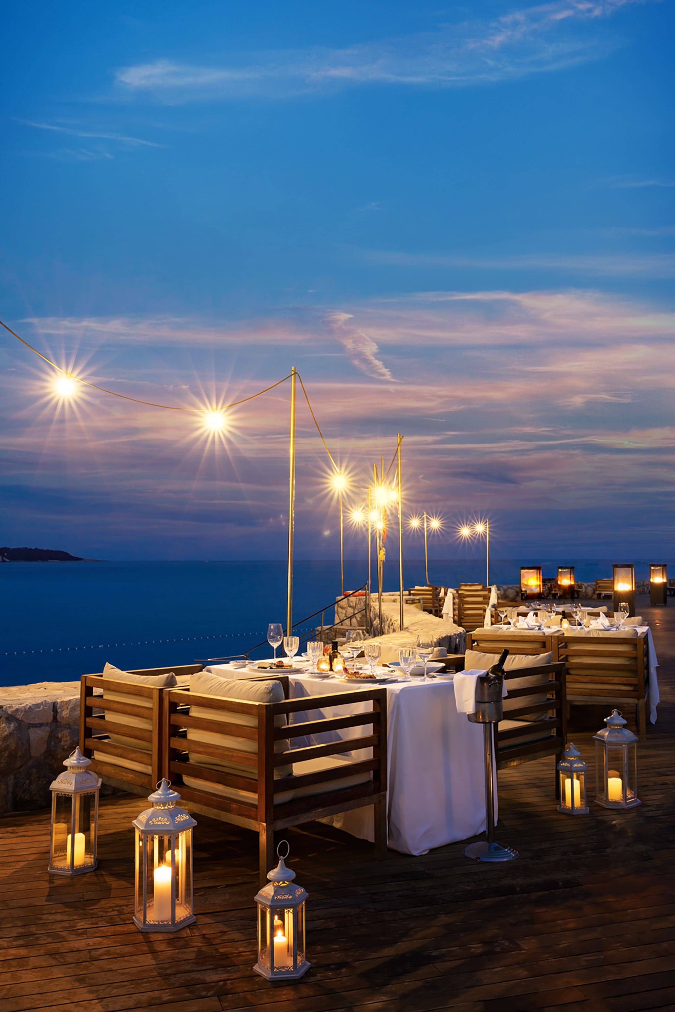 A stunning vertical shot of an outdoor dining setup at the Libertas Fish Restaurant at Rixos Premium Dubrovnik at twilight. Tables are set with white tablecloths and fine dining place settings on a wooden deck, with seating made of wooden slats. The area is lit by strings of warm overhead lights and several white decorative lanterns with lit candles on the deck. The setting overlooks the deep blue Adriatic Sea under a vibrant blue and purple-streaked sky.