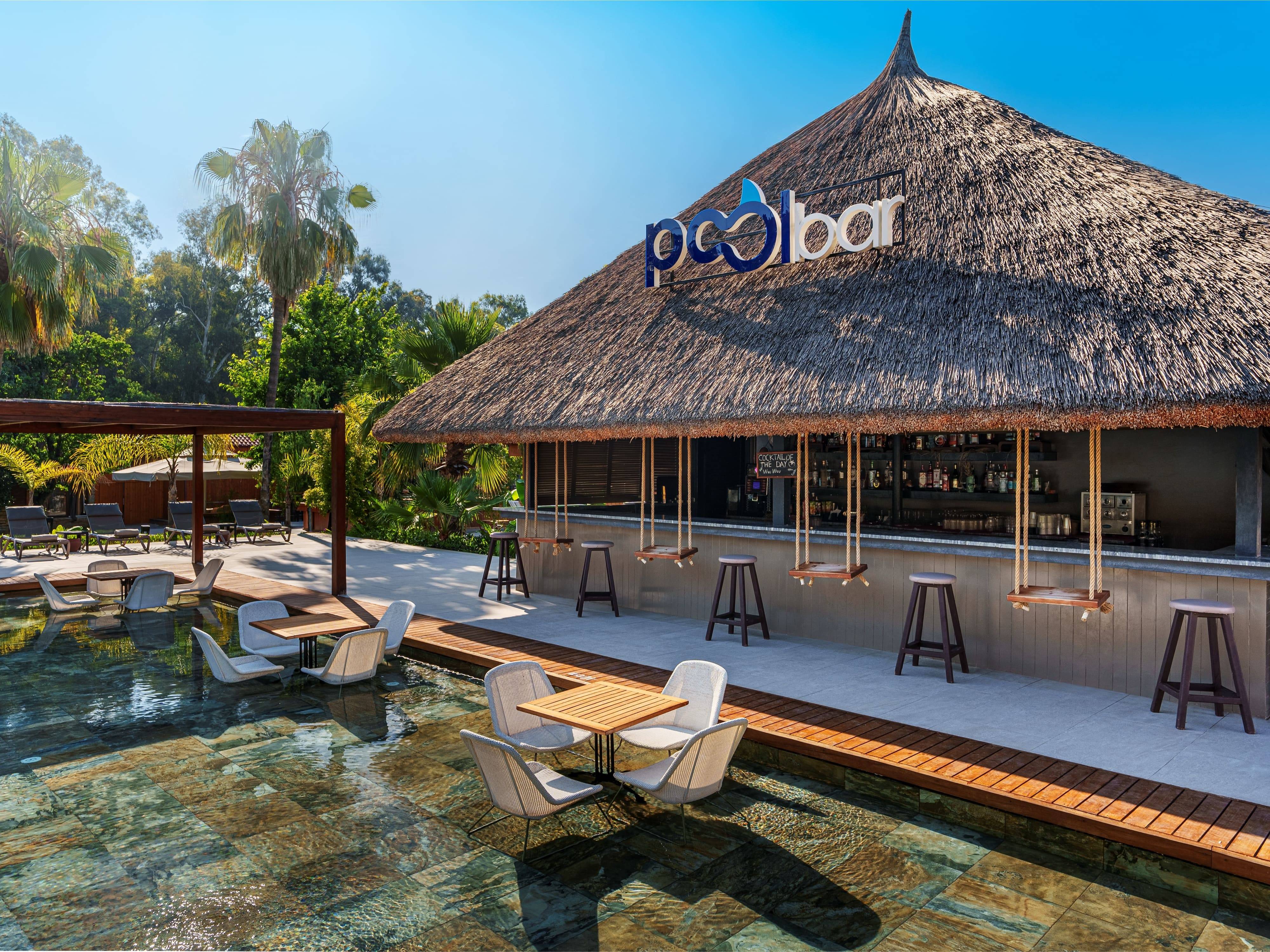Thatched-roof Pool Bar at Rixos Göcek surrounded by lush greenery, featuring in-water seating, wooden decks, and hanging swing stools under a tropical-style canopy.