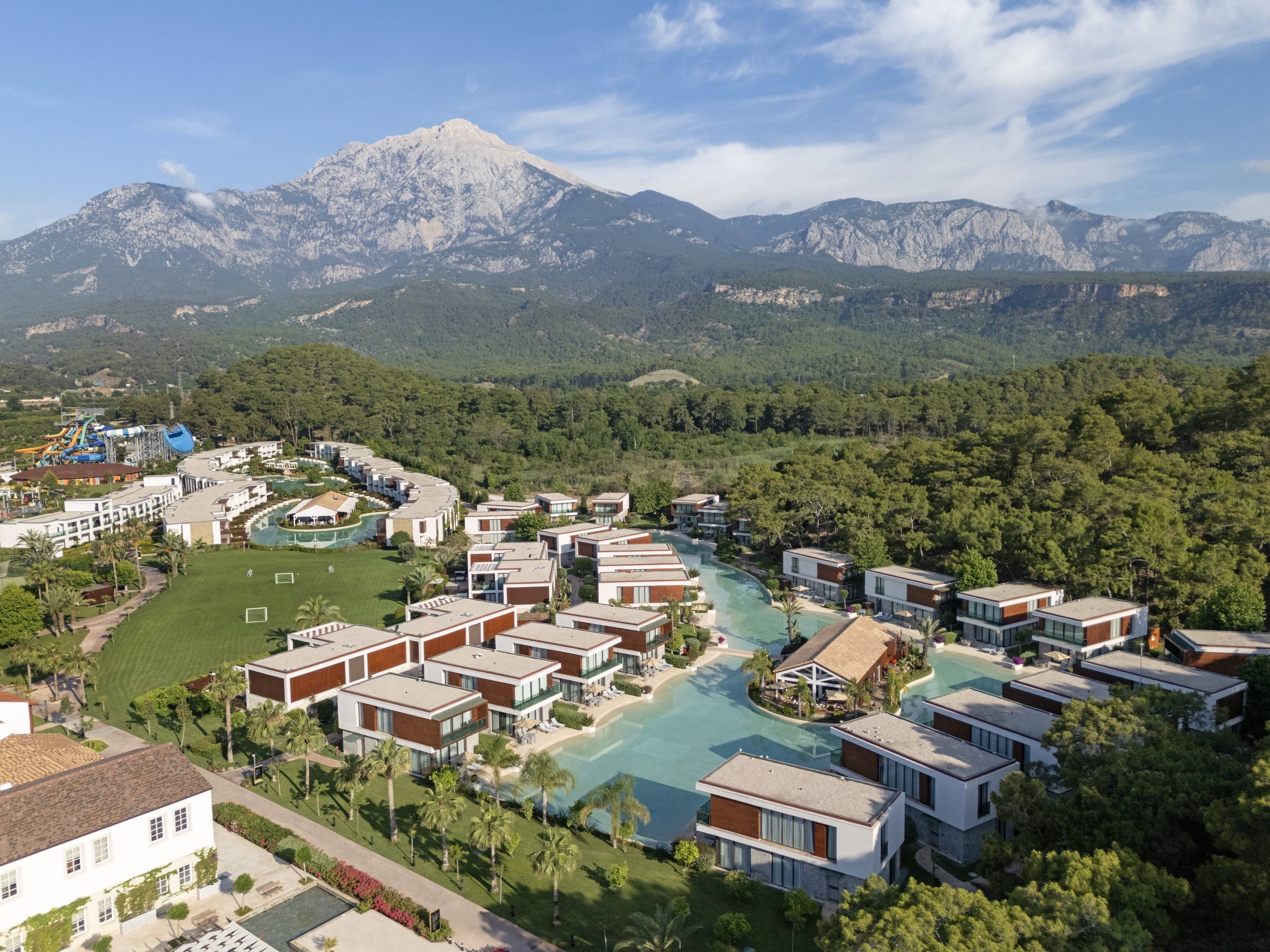 Aerial view of Rixos Premium Tekirova, a modern resort with villas surrounded by a lagoon, lush greenery, and mountains in the background under a blue sky with scattered clouds.